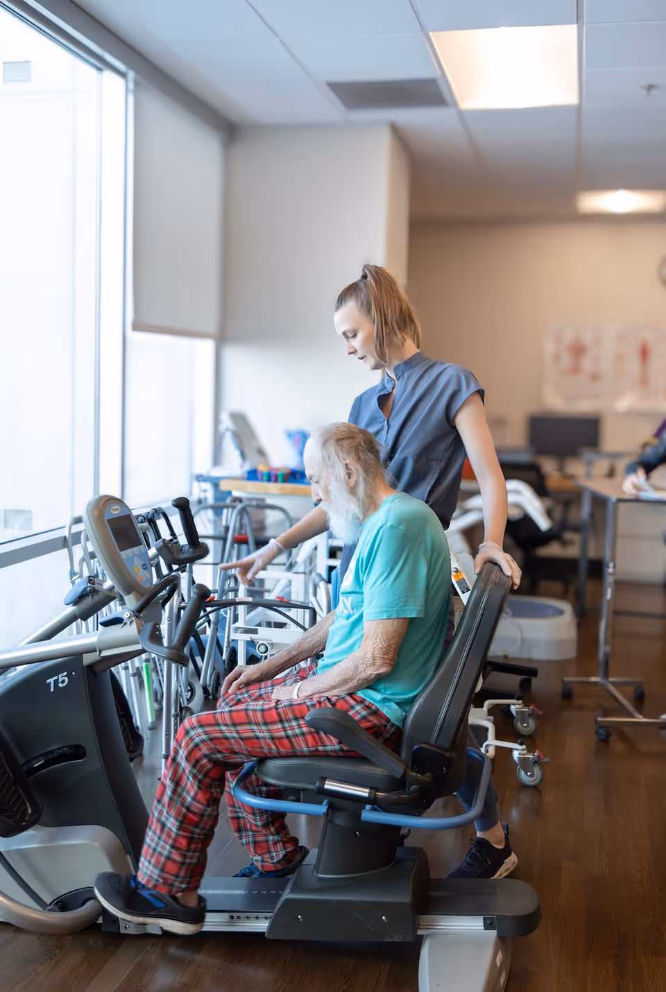 An elderly man in plaid pants and a teal shirt is seated on a recumbent exercise bike while a young female caregiver or physical therapist stands beside him, pointing at the bike's controls in a bright room with large windows and exercise equipment.