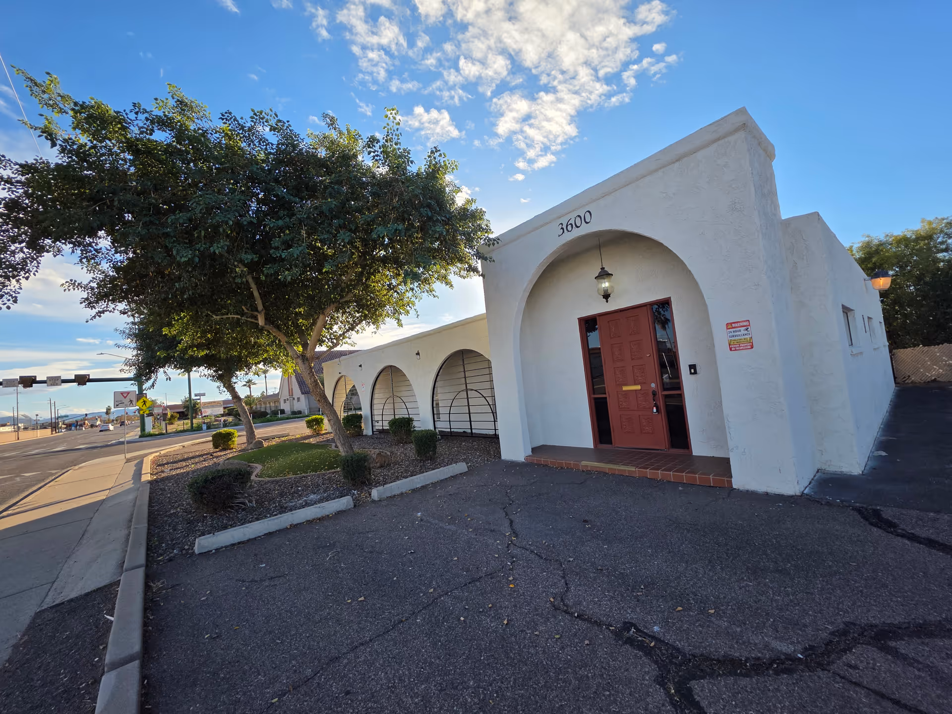 White stucco building with arched entrance and red door, set beside a sidewalk under blue sky.