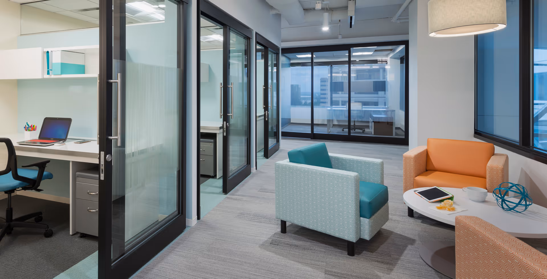 Modern office-like lounge with glass doors, desks, and colorful seating in a senior care facility.