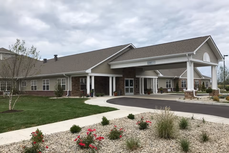Exterior view of a single-story senior living facility building with beige siding, stone accents, and a covered entrance driveway. The foreground features a landscaped area with rocks, small bushes, and red flowers. The sky is cloudy.