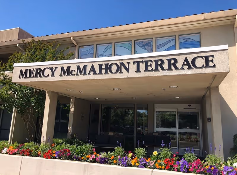 Entrance of Mercy McMahon Terrace facility with a covered walkway, large sign displaying the facility name, colorful flower beds in front, and glass doors leading inside.