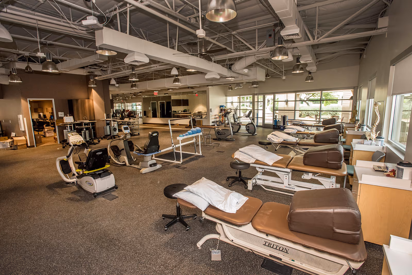 Spacious therapy gym with exercise equipment, treatment tables, and large windows.
