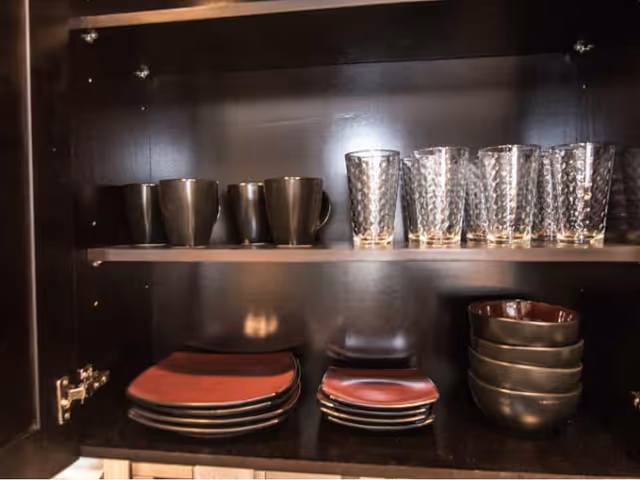 Open kitchen cabinet with neatly arranged black mugs, clear textured drinking glasses, black bowls, and black plates with red centers.