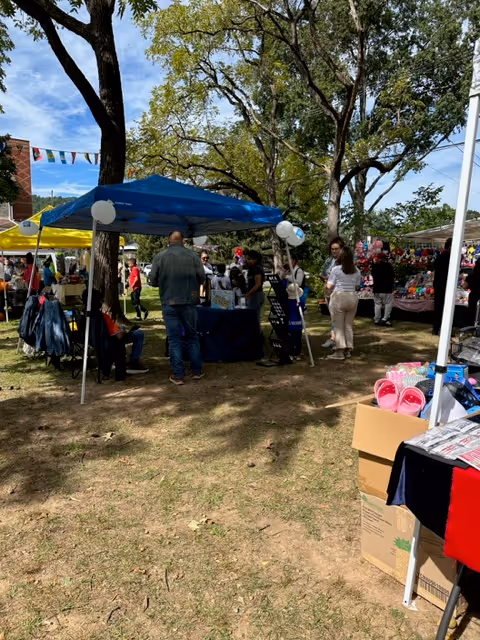 Outdoor community market with tents, balloons, and people shopping under trees.