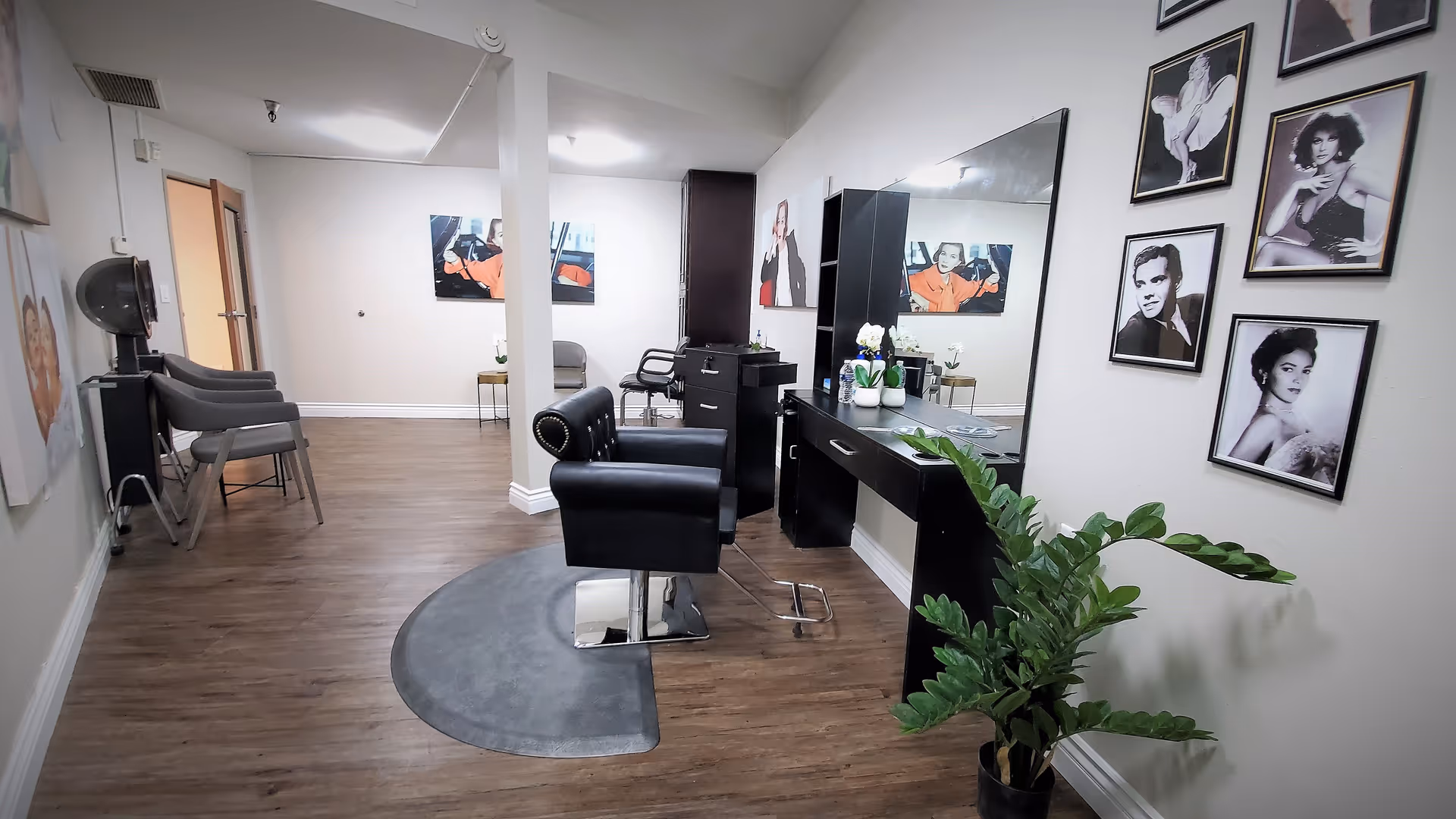 Interior view of a salon area in Terraza Court Senior Living featuring a black salon chair on a gray mat in front of a large mirror with a black counter. The walls are decorated with framed black and white portraits and colorful artwork. There are additional chairs and a hair dryer in the background, along with a potted plant near the mirror.
