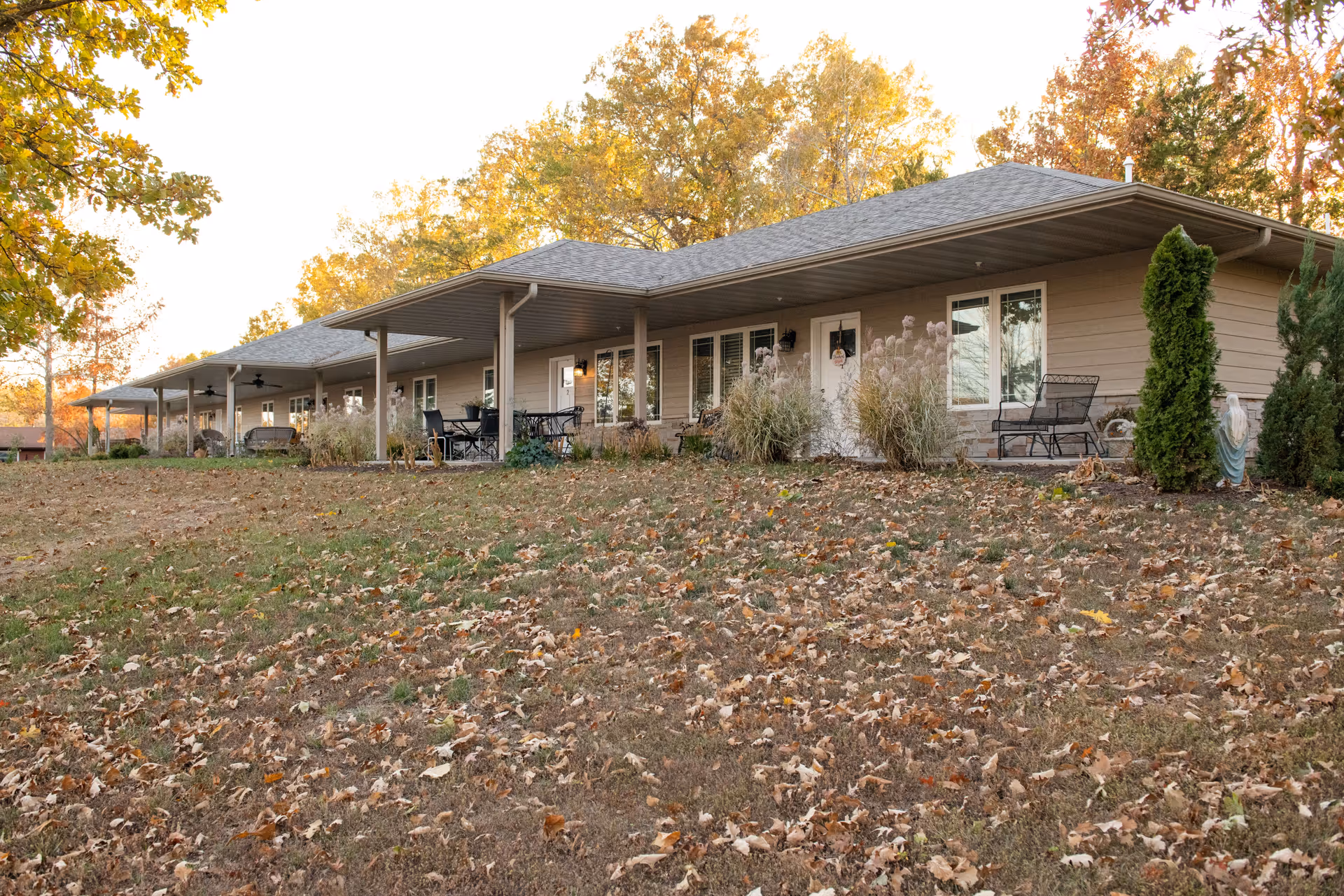 Single-story senior living building with a covered porch, outdoor seating, and autumn trees and lawn in front.