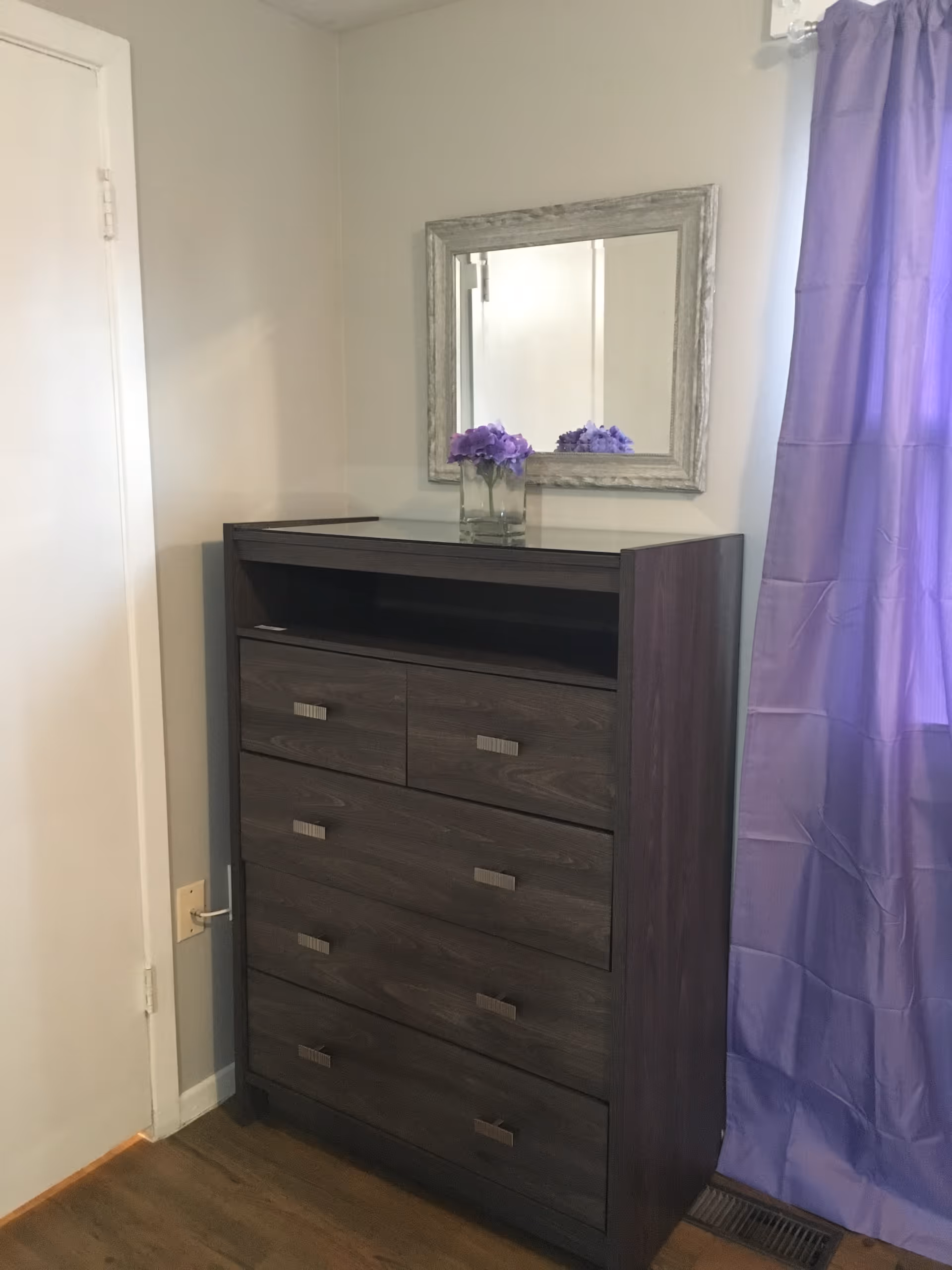 A dark wooden dresser with multiple drawers stands against a light-colored wall. Above the dresser is a rectangular mirror with a rustic wooden frame. On top of the dresser is a clear glass vase containing purple flowers. To the right, a purple curtain hangs beside a window, and to the left is a closed white door. The floor is wooden.