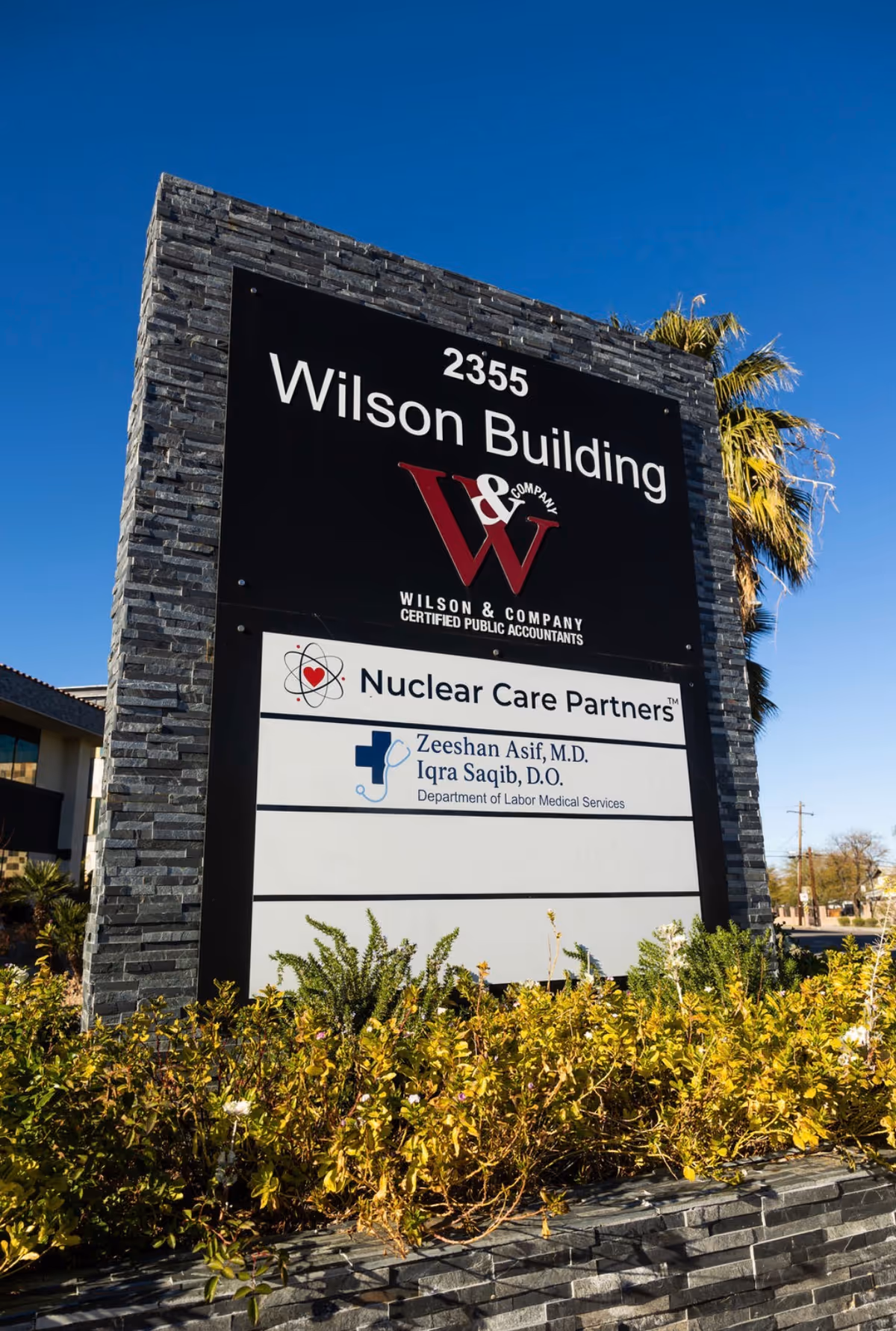 Tall stone sign displaying Wilson Building and Nuclear Care Partners logos under a blue sky.