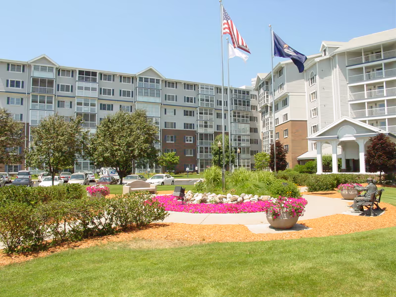 Landscaped front courtyard and entrance of a multi-story senior living building with flags and flower beds.