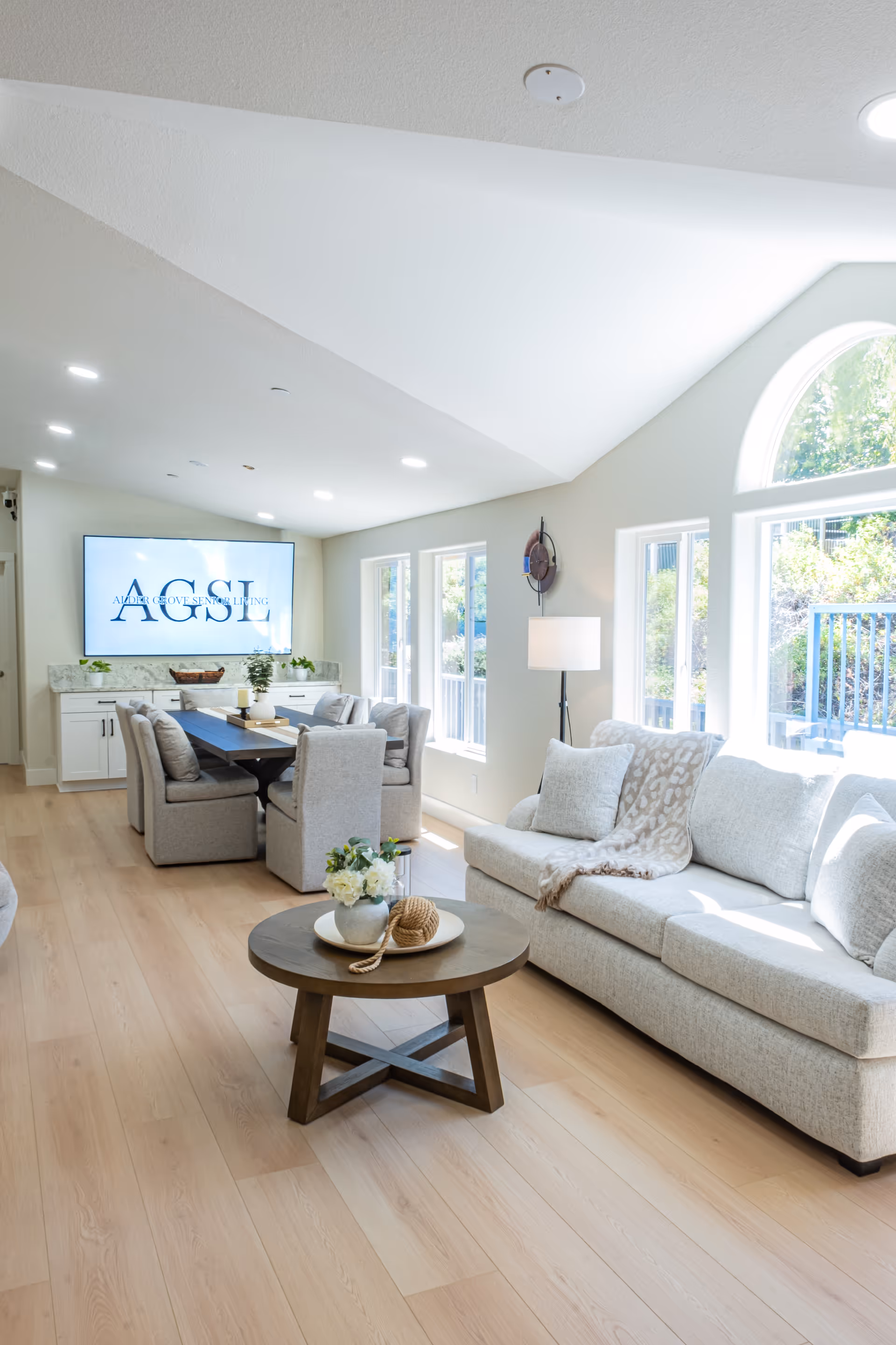 Bright senior living common area with beige sofa, round coffee table, and dining seating.