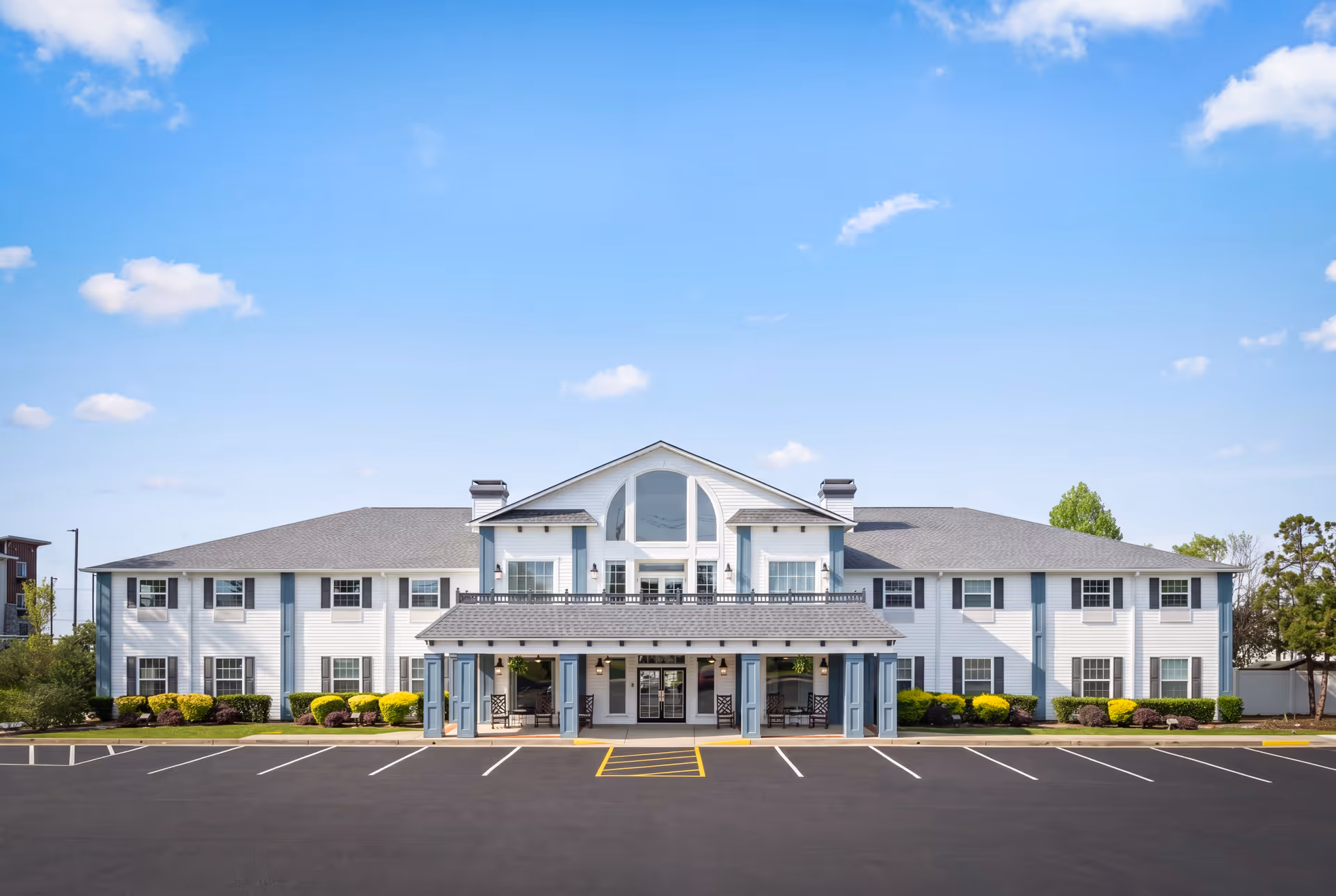 Two-story light-blue senior living building with central entrance and empty parking lot.