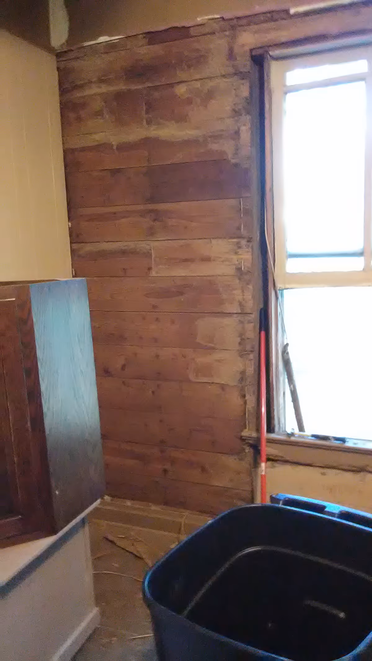 Interior renovation scene with exposed wooden wall, window, and blue bin in an unfinished room.