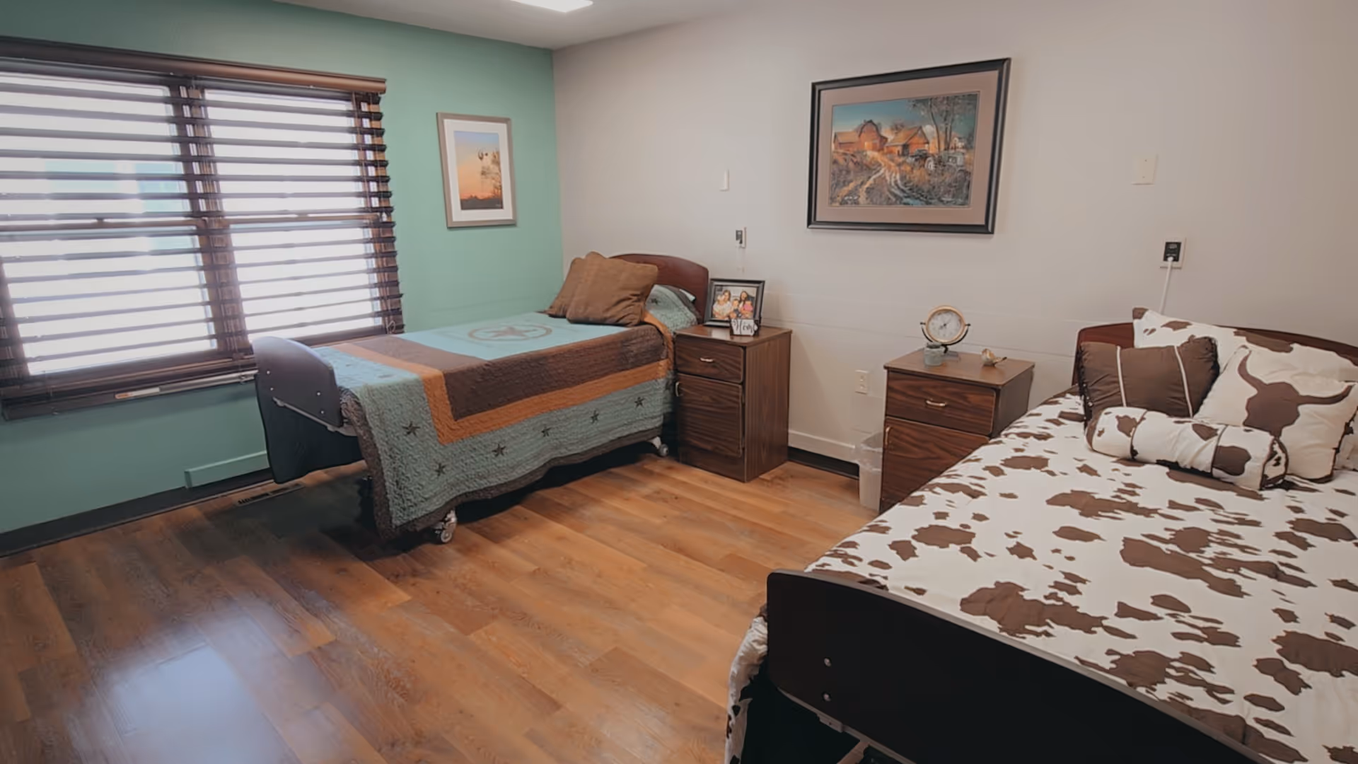 Two beds in a shared senior living bedroom with nightstands, artwork, and wooden flooring.