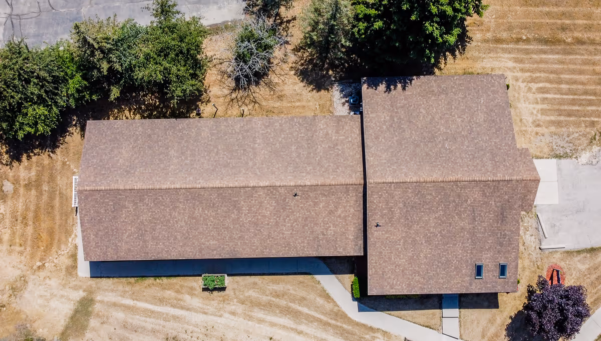 Aerial view of a single-story building with a brown shingled roof surrounded by dry grass and a few trees, showing a concrete walkway leading to the entrance.