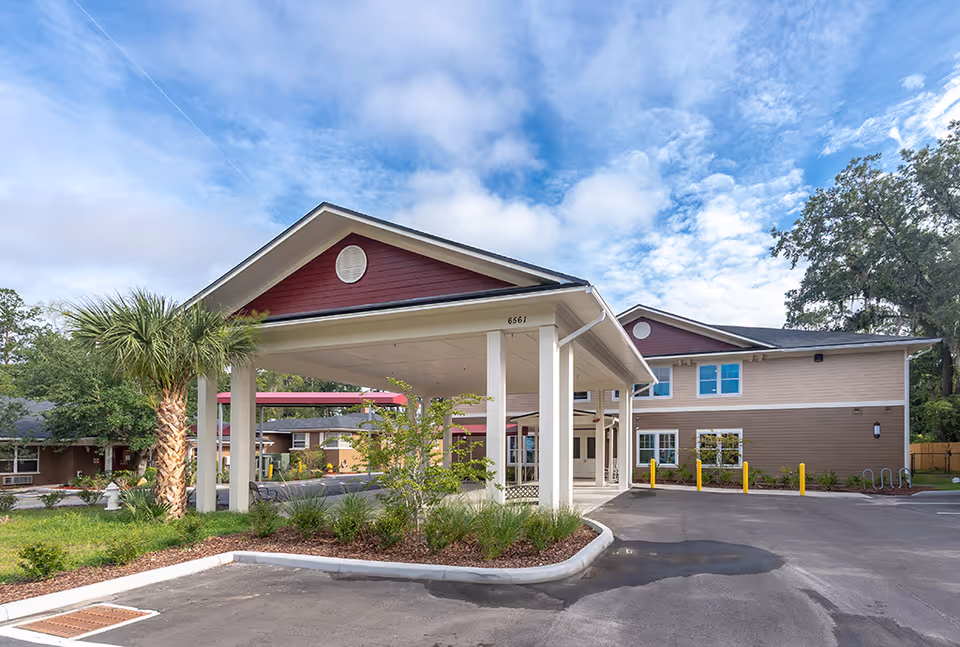 Front entrance and porte-cochere of a two-story assisted living building with palm trees and a parking area under a partly cloudy sky.