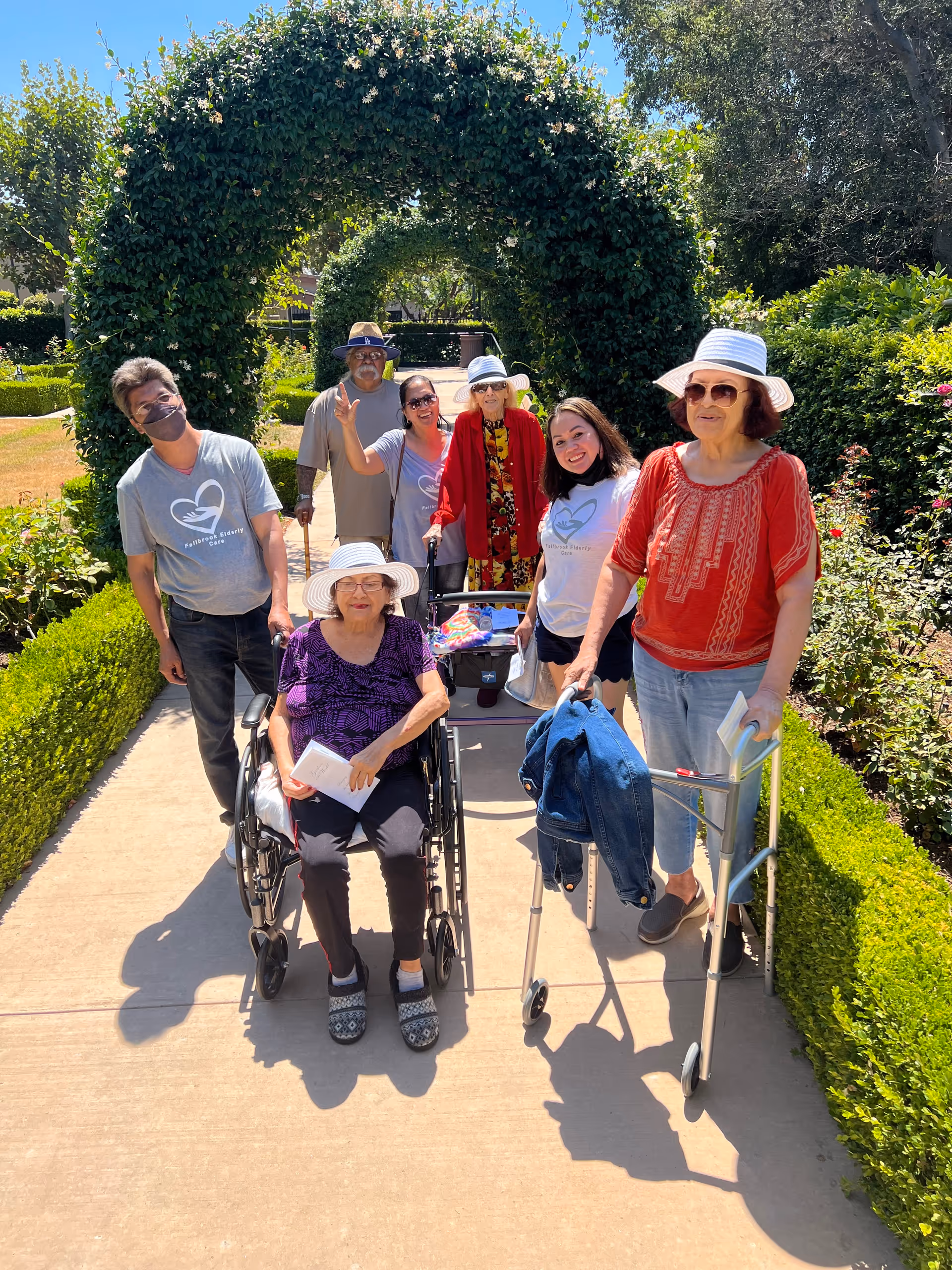 A group of elderly and middle-aged people enjoying a sunny day outdoors in a garden with green hedges and archways covered in foliage. Two elderly women are using mobility aids, one in a wheelchair and another with a walker. Some people are wearing hats and sunglasses, and a few are wearing matching gray t-shirts with a heart logo and the text 'Fallbrook Elderly Care'.