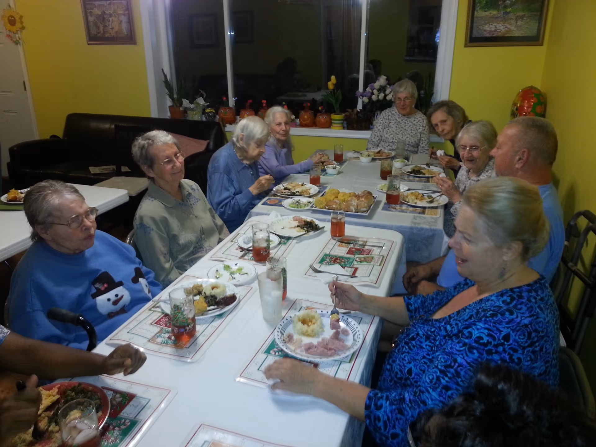 A group of elderly people sitting around a long dining table enjoying a meal together in a room with yellow walls and a window decorated with plants and small pumpkin decorations.