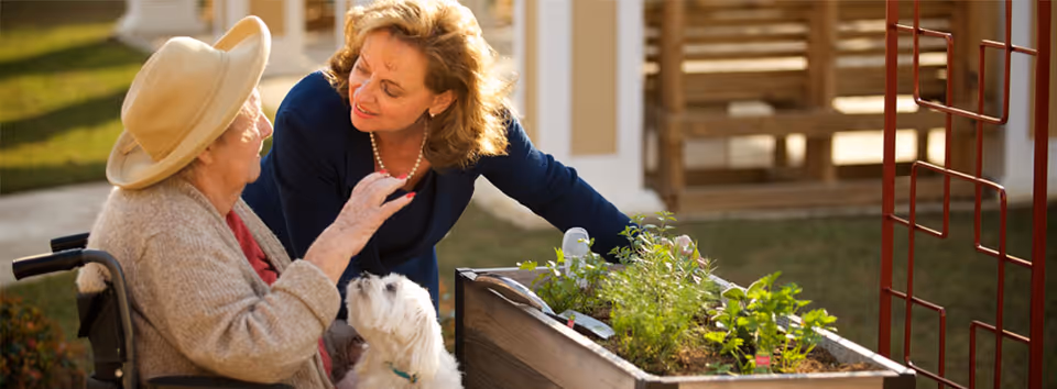 An elderly woman in a wheelchair wearing a hat and a younger woman leaning towards her, both smiling and interacting outdoors near a raised garden bed with plants and a small white dog sitting on the elderly woman's lap.