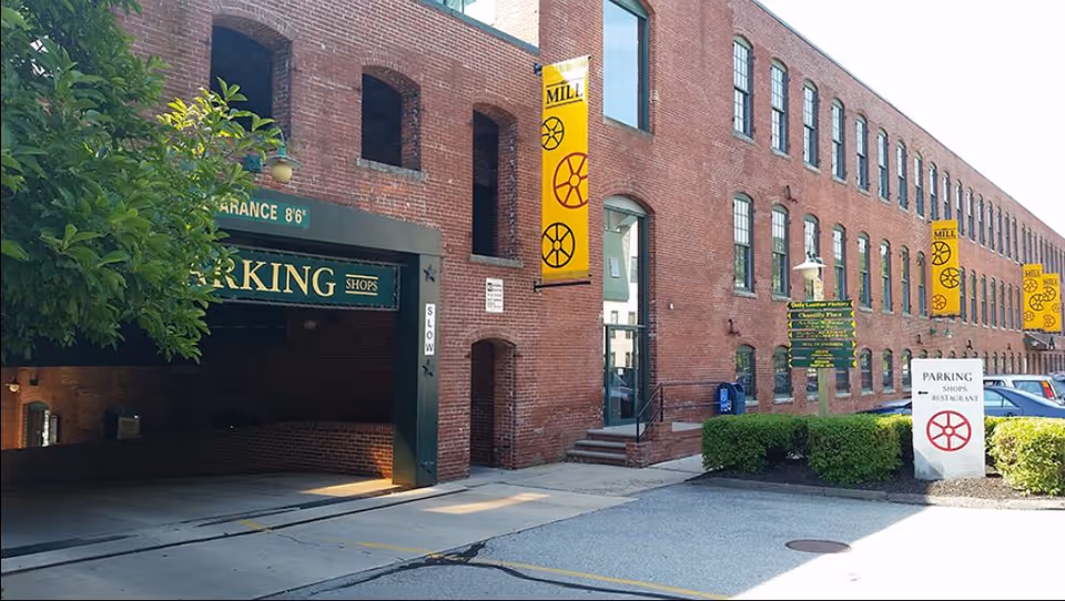 Brick, multi-story facility exterior with yellow banners, a parking entrance, and trimmed hedges along the sidewalk.