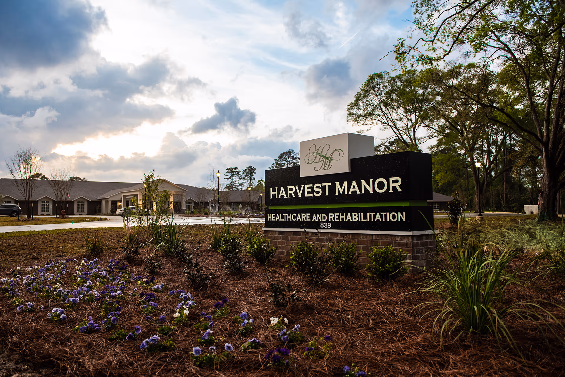 Entrance sign reading 'Harvest Manor Healthcare and Rehabilitation' with landscaped flowers and the facility building under a cloudy sky.