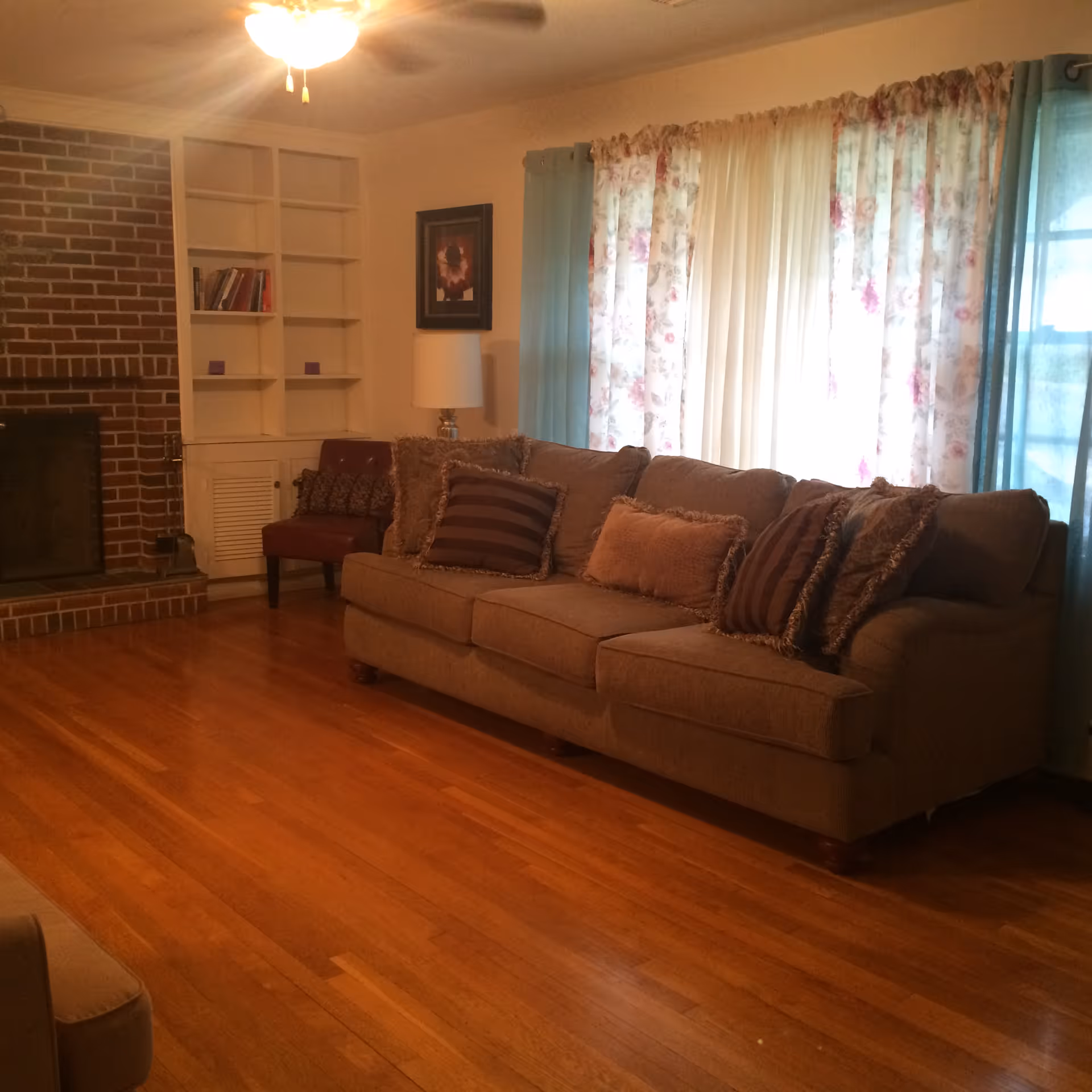 A cozy living room with a brown sofa adorned with multiple cushions, a red armchair, a white built-in bookshelf, a brick fireplace, a lamp, and floral curtains covering a window.