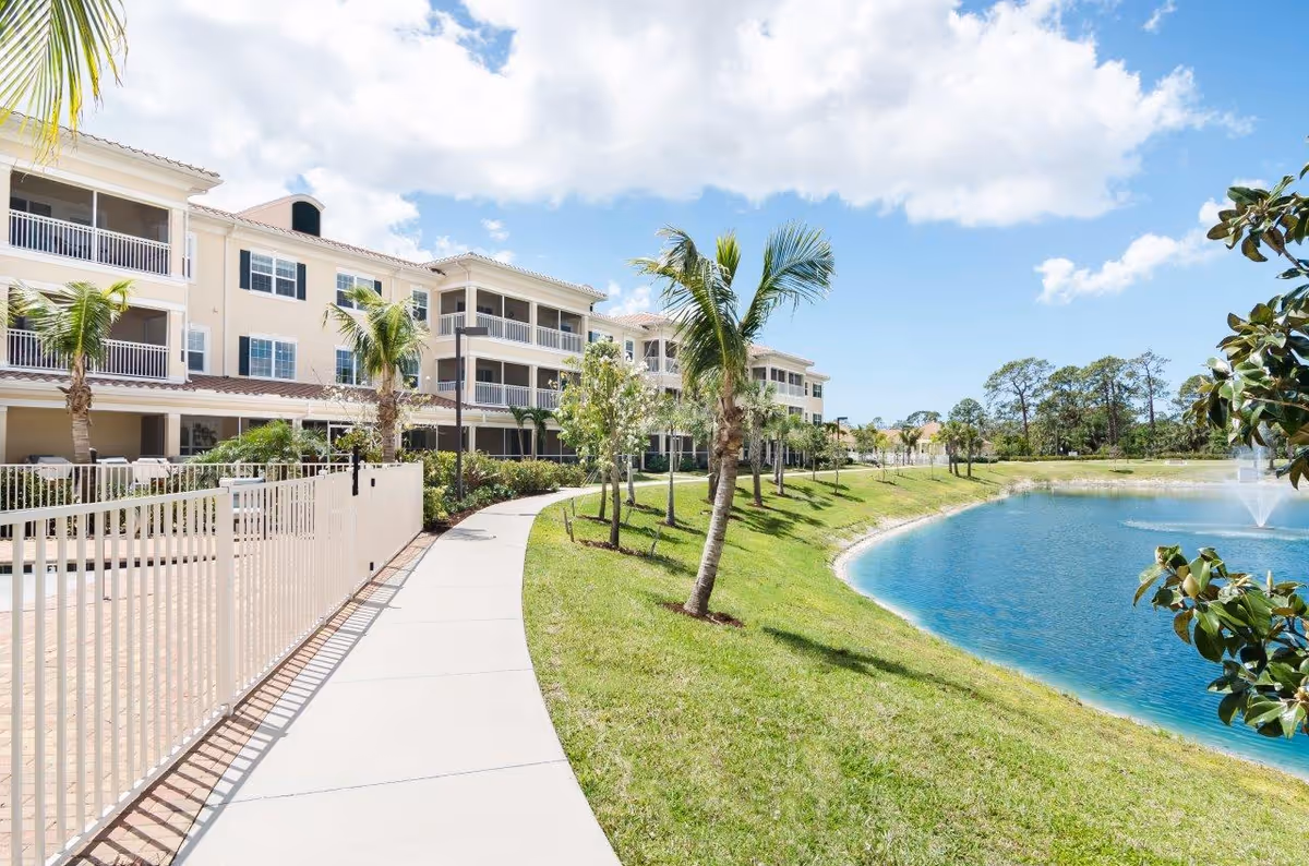 Sunlit exterior view of American House Bonita Springs III with a pond and walkway.