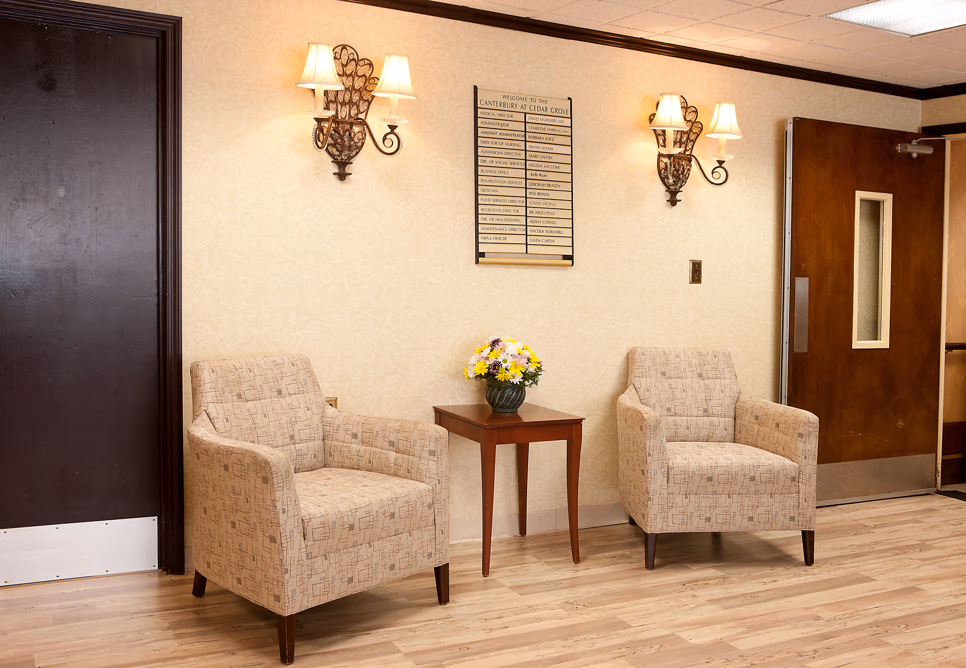 Two beige armchairs sit with a small table and flowers in a waiting area.