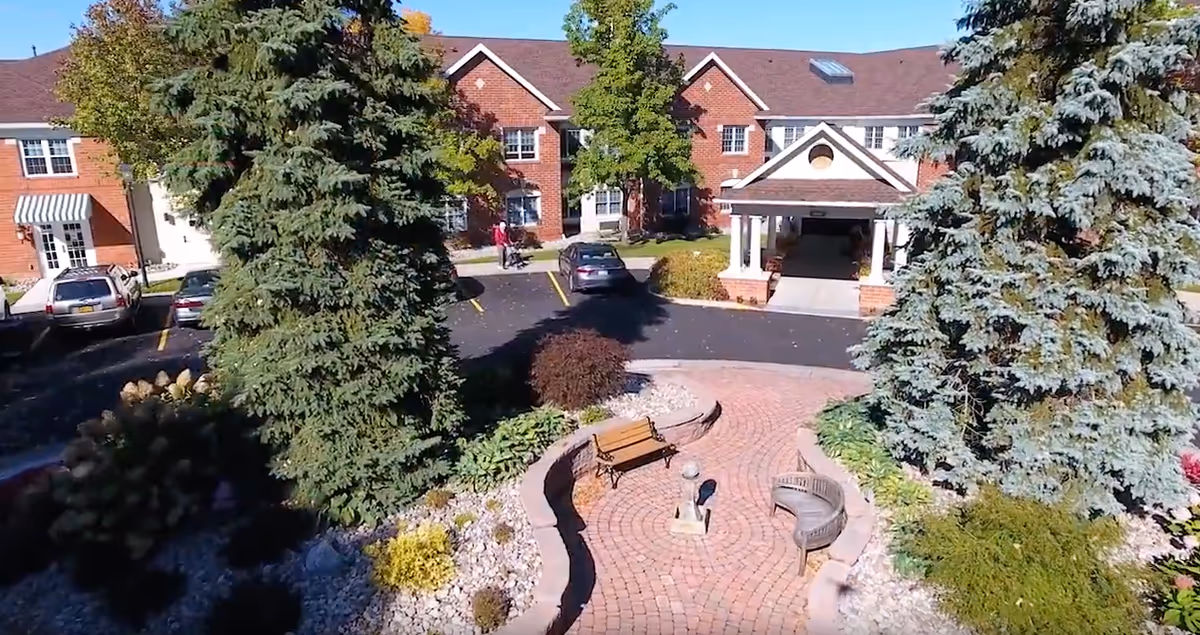 Brick senior living building entrance with a circular paved courtyard, benches, large evergreen trees and parked cars.