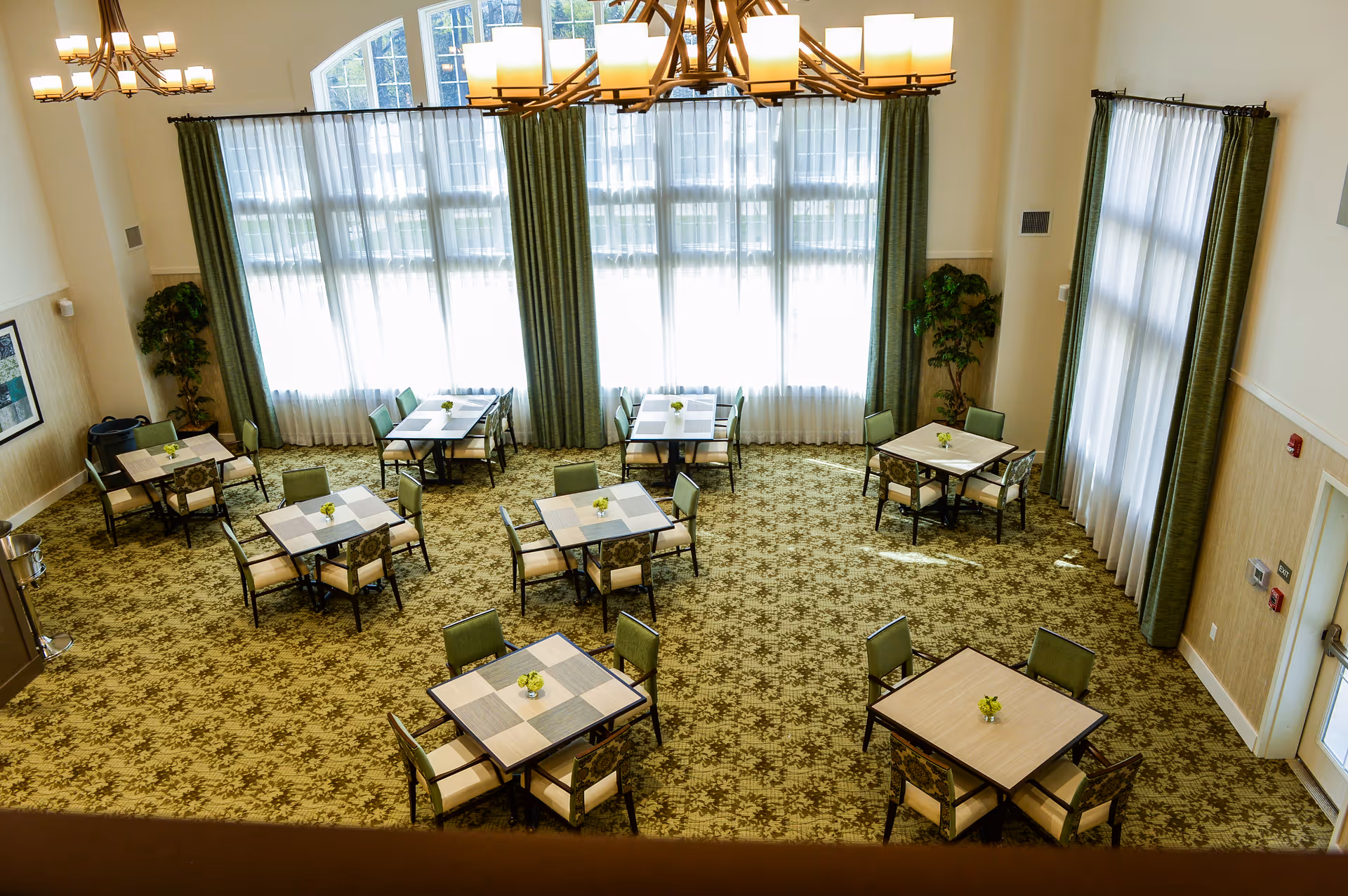 A spacious dining room with several square tables, each surrounded by four chairs. The room features large windows with sheer white curtains and green drapes, patterned carpet, and two chandeliers hanging from the ceiling. Small flower arrangements are placed on each table.