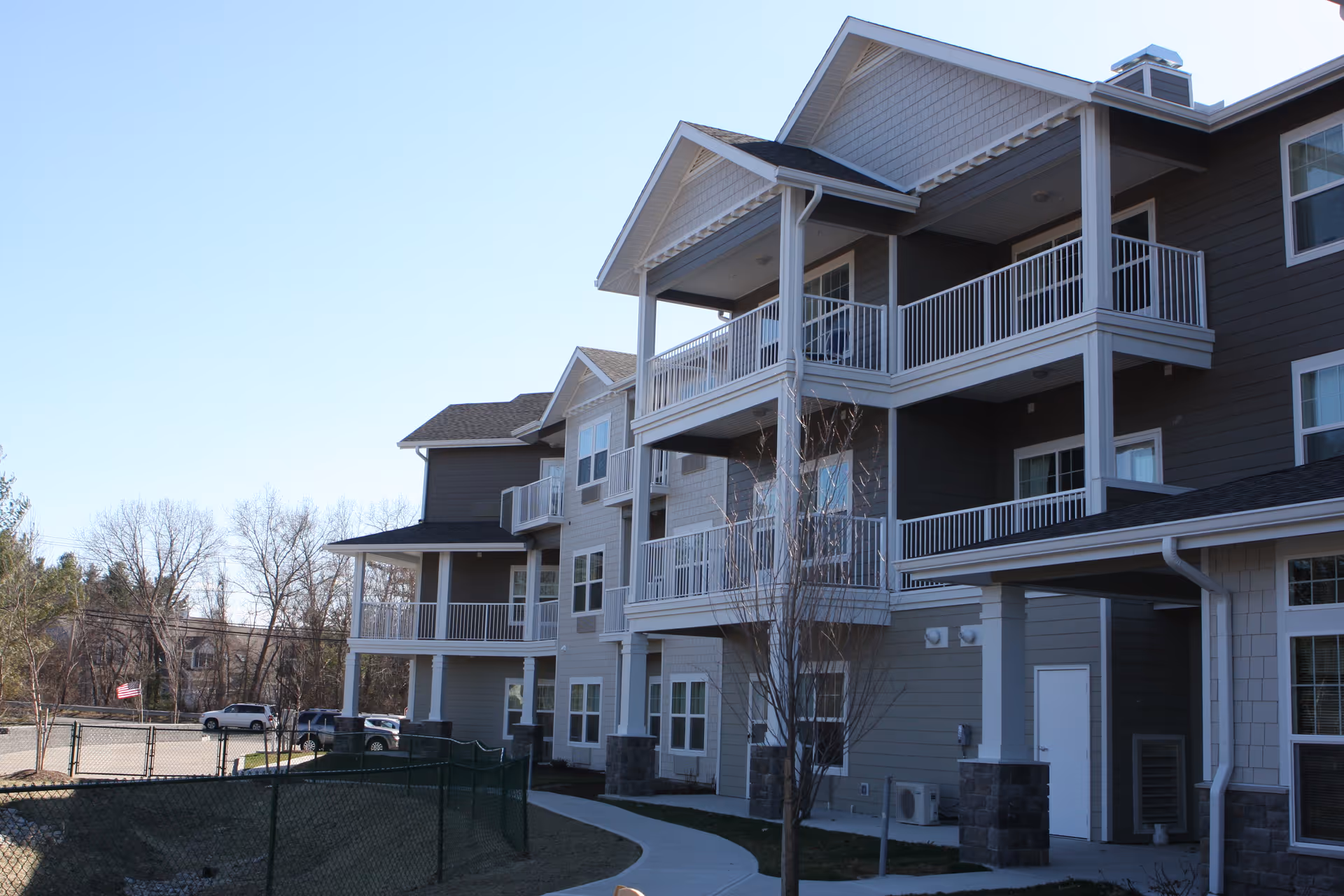 Exterior view of a multi-story residential building with balconies, a paved walkway, and some trees in front. The building has gray siding with white trim and railings, and the sky is clear and blue.