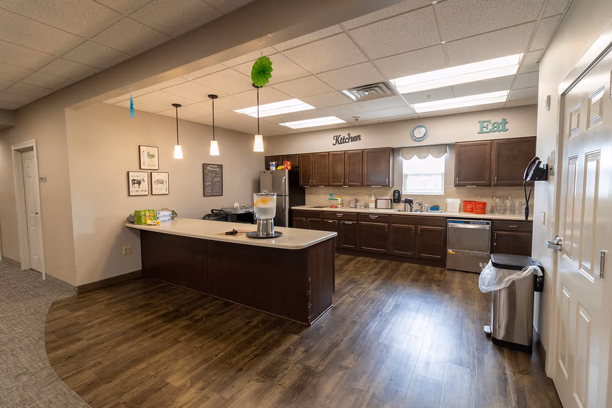 A spacious kitchen area with dark wooden cabinets, a countertop island with a water dispenser, and stainless steel appliances including a refrigerator and dishwasher. The room has wood flooring, overhead fluorescent lighting, and wall decorations including the words 'Kitchen' and 'Eat'.