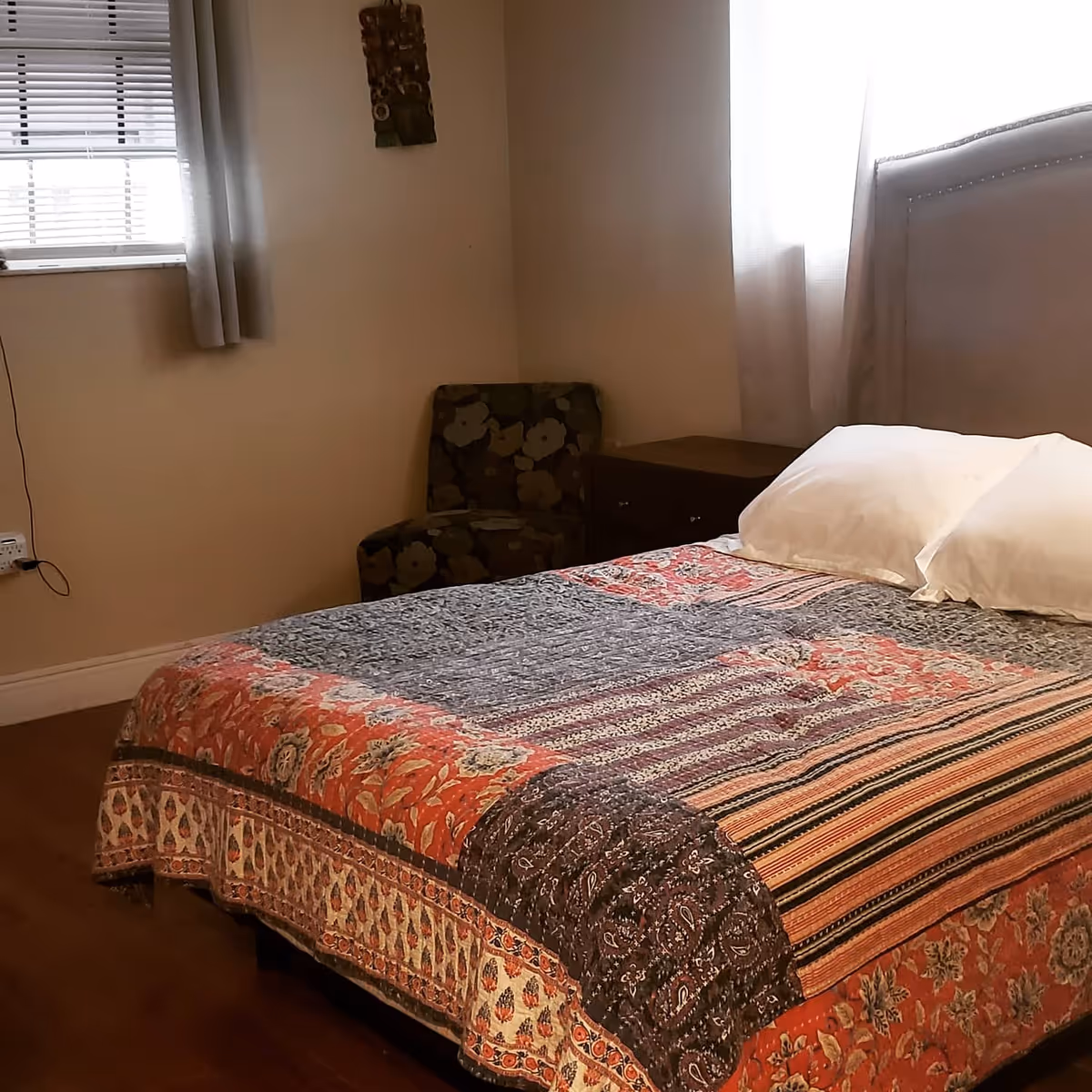 A bedroom with a bed covered in a colorful patterned quilt and two white pillows. There is a floral-patterned armchair in the corner next to a wooden nightstand. Two windows with white curtains allow natural light into the room.