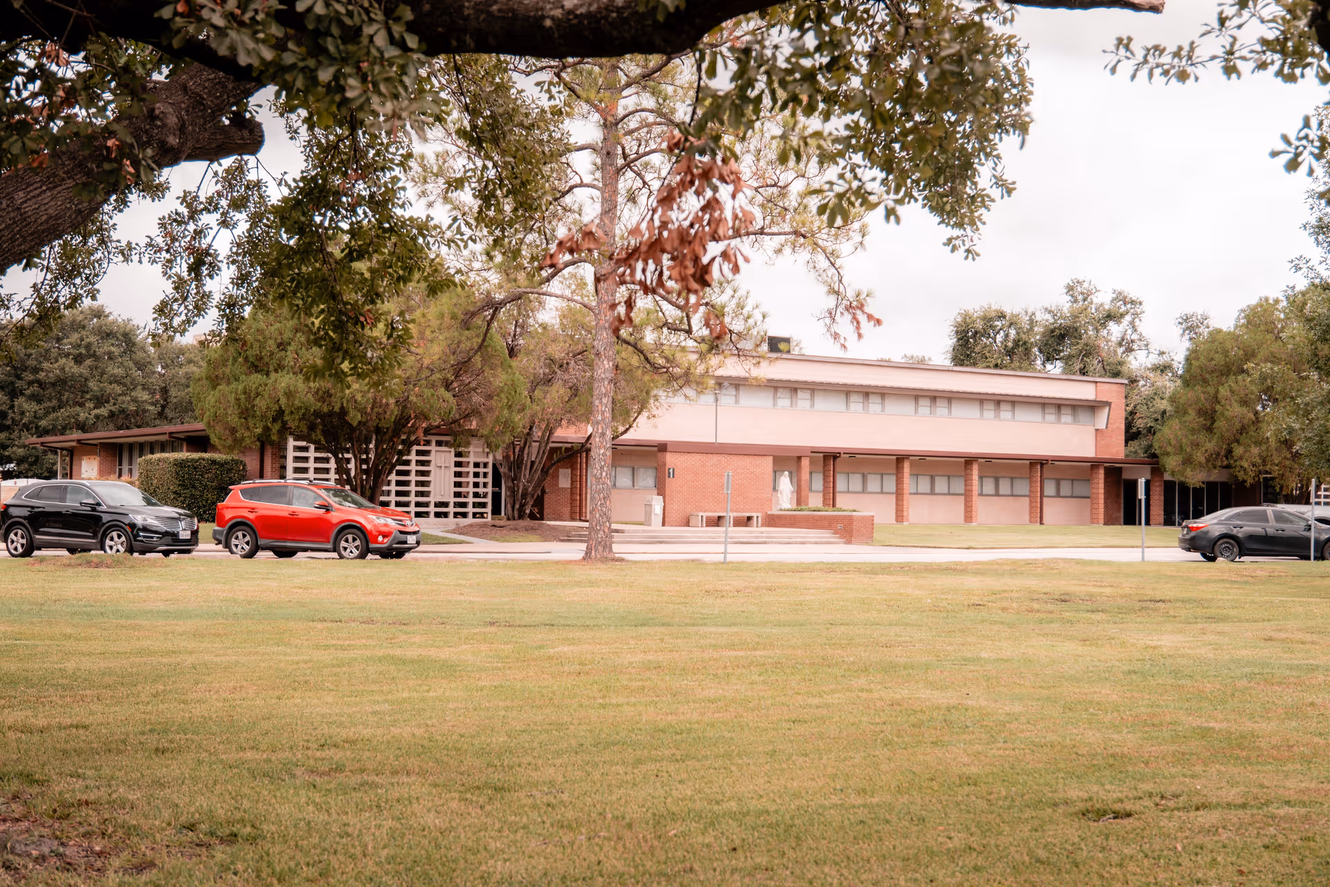 Brick two-story building set behind a grassy lawn and trees with several cars parked along the driveway.