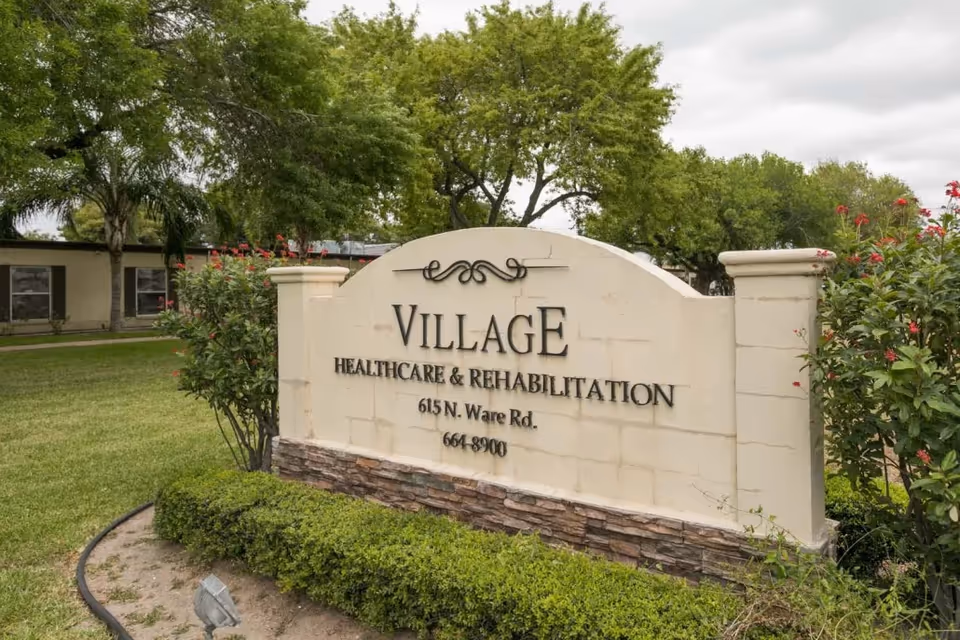 A beige stone sign reading Village Healthcare & Rehabilitation stands at a landscaped entrance.