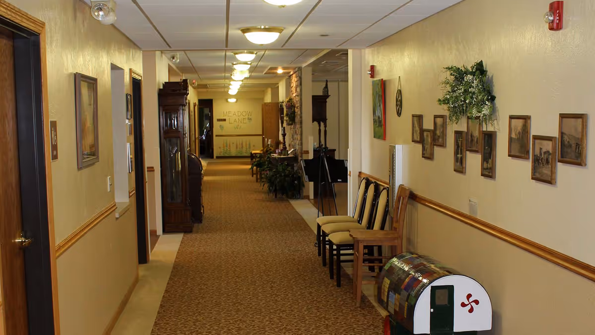 Carpeted interior hallway of a senior living facility with chairs, framed pictures, plants, and a decorative mailbox, with a 'Meadow Lane' sign visible at the far end.