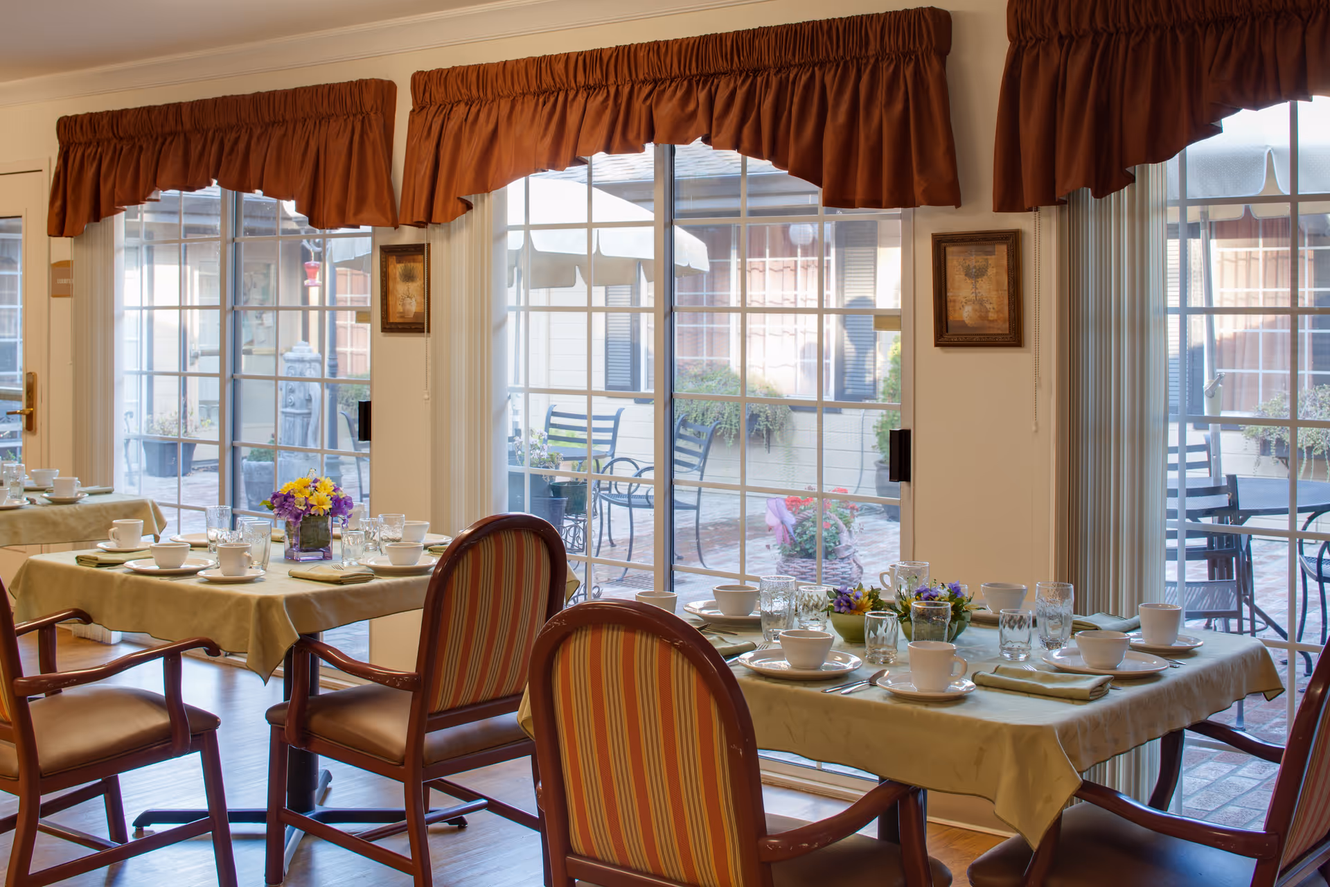 A dining area with tables set for a meal, including plates, bowls, glasses, and napkins. The tables have beige tablecloths and small flower arrangements. The room has large sliding glass doors with brown valance curtains, leading to an outdoor patio with chairs and tables.