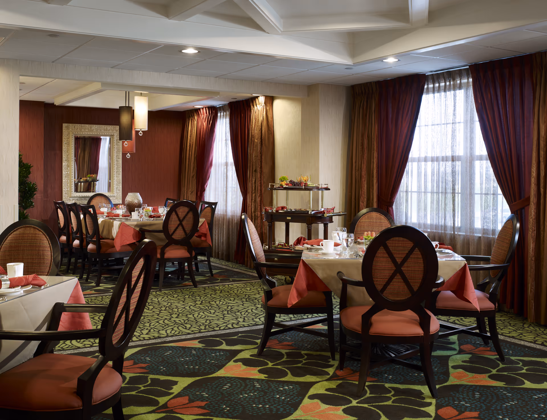 Senior living dining room with round tables set for meals, burgundy curtains, and warm lighting.