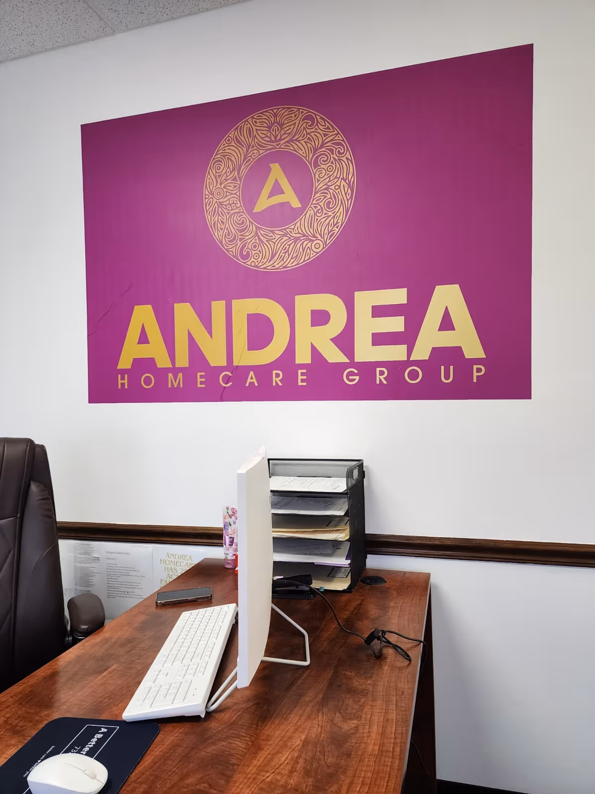 Office desk with computer, filing rack, and a purple Andrea Homecare Group sign on wall.
