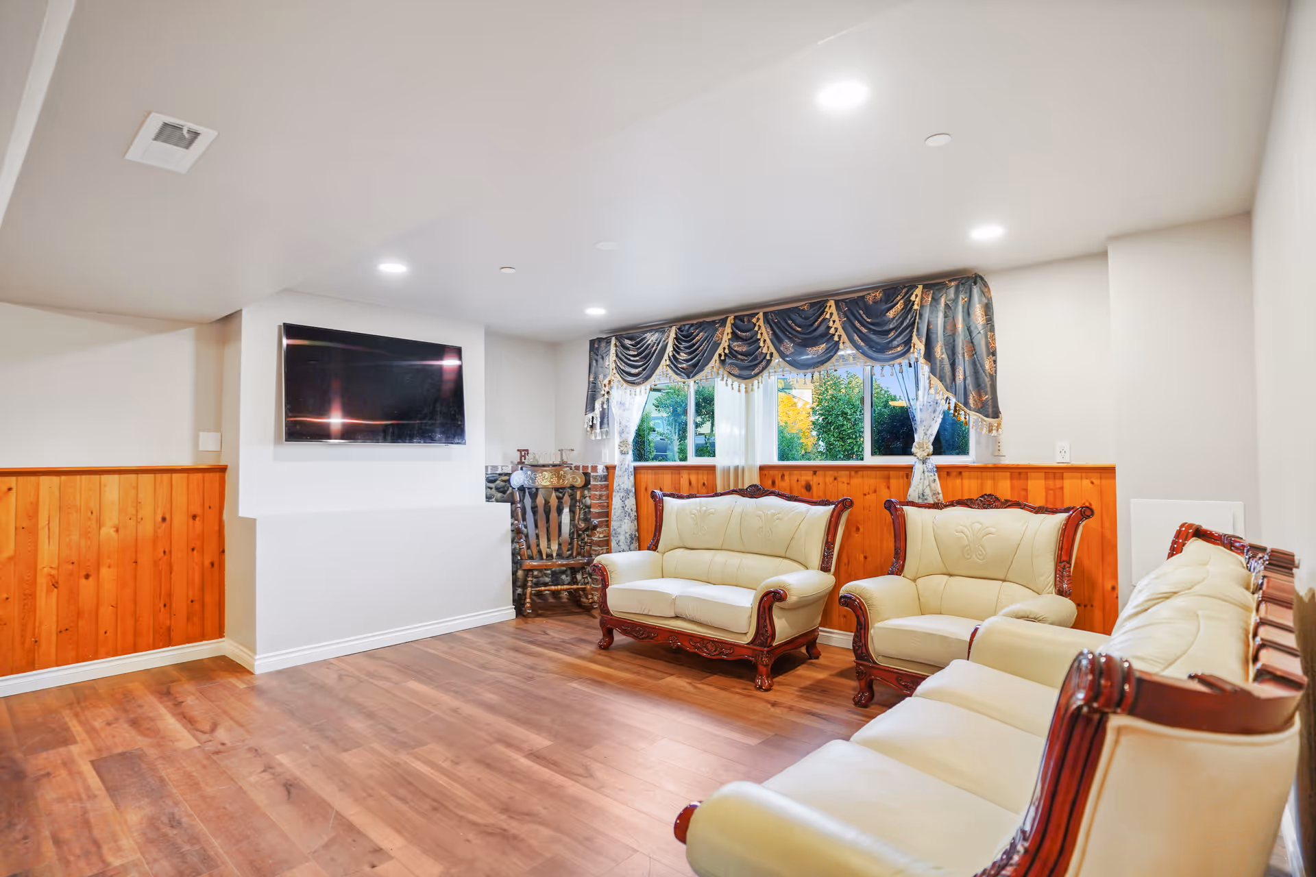 A bright living room with wooden flooring and wood paneling on the lower half of the walls. The room features cream-colored leather sofas with dark wooden frames arranged around the space. A flat-screen TV is mounted on a white wall, and a window with decorative blue and gold curtains lets in natural light. There is also a wooden rocking chair near the window.