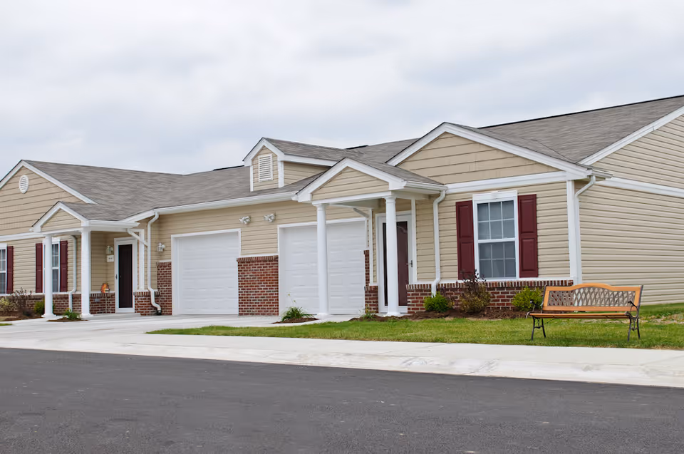 Front exterior view of a beige senior living facility with garage doors and a bench.