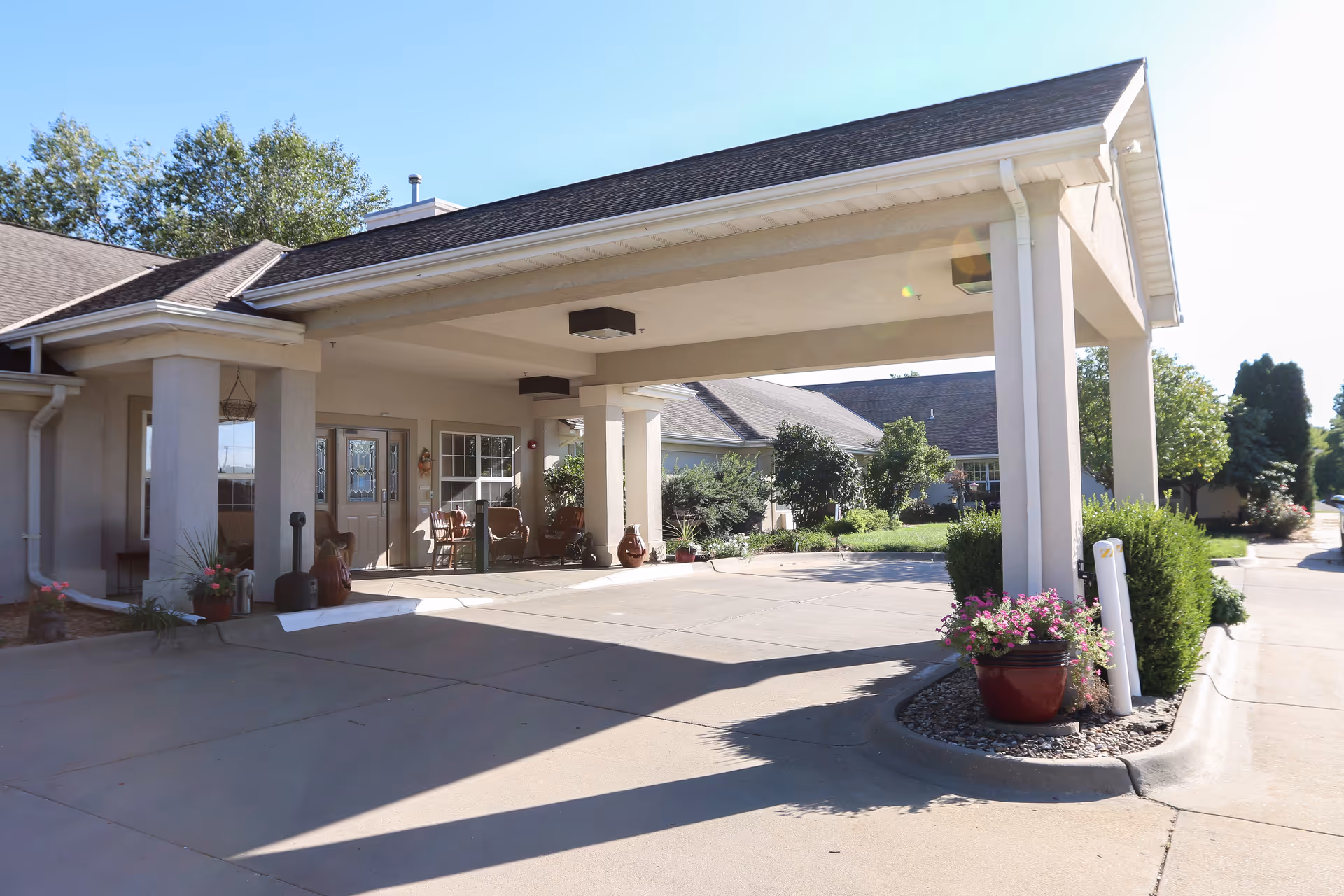 Senior living facility entrance with a covered driveway, seating, and potted plants.