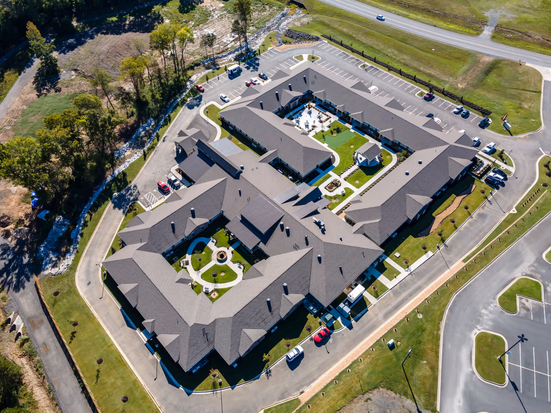 Aerial view of Crestview Senior Living facility showing a large, U-shaped building with multiple wings surrounding landscaped courtyards. The building is surrounded by parking lots, roads, and green spaces with trees and grass.
