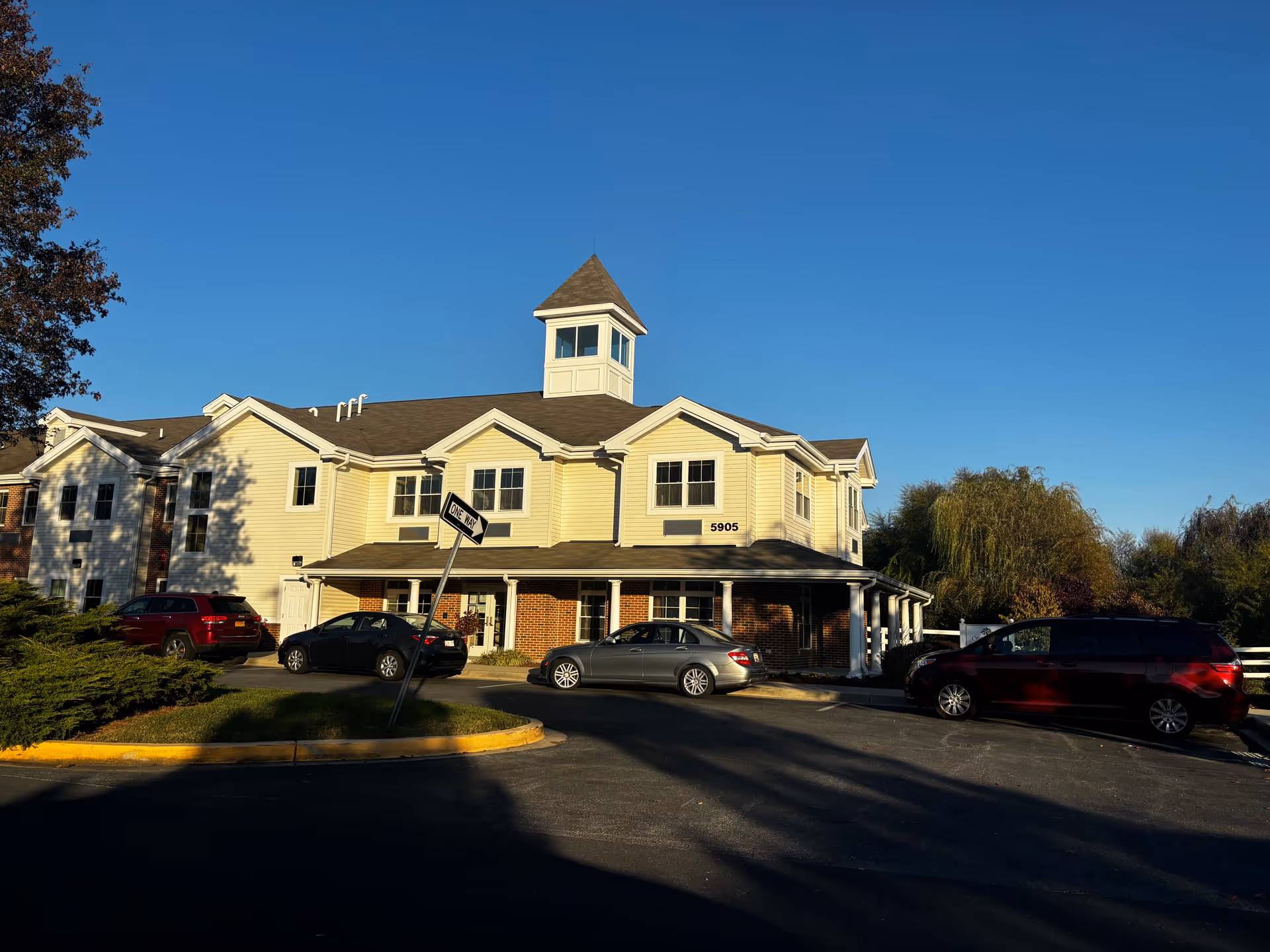 Front exterior of a two-story senior living building with a cupola, cars parked at the entrance, and a clear blue sky.