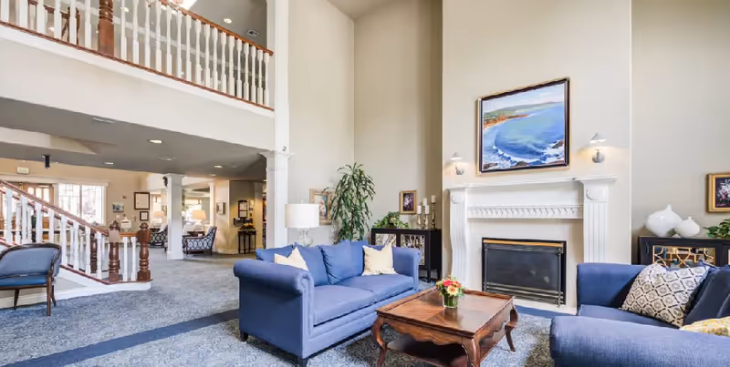 Spacious senior living lobby lounge with blue sofas, fireplace, and staircase balcony.