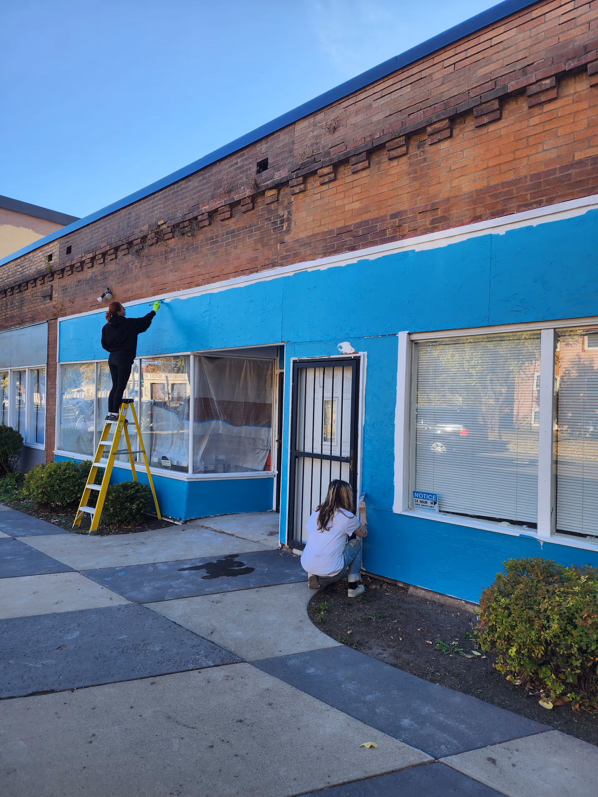 Two people repaint the facility's blue exterior wall with a ladder and painting supplies.