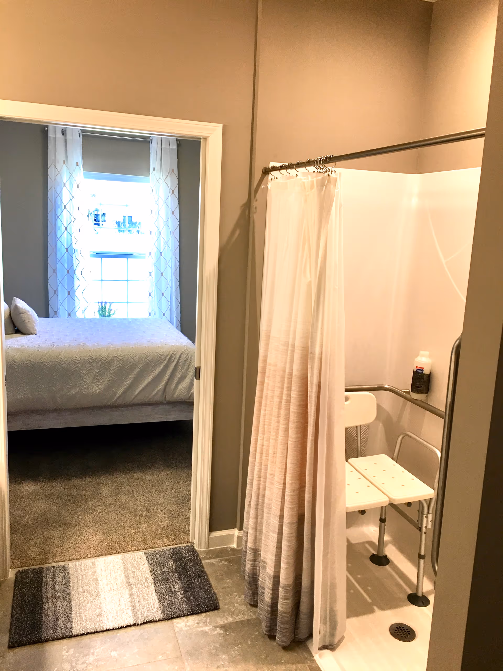 Accessible bathroom with shower chair and grab bars; bedroom visible through doorway.