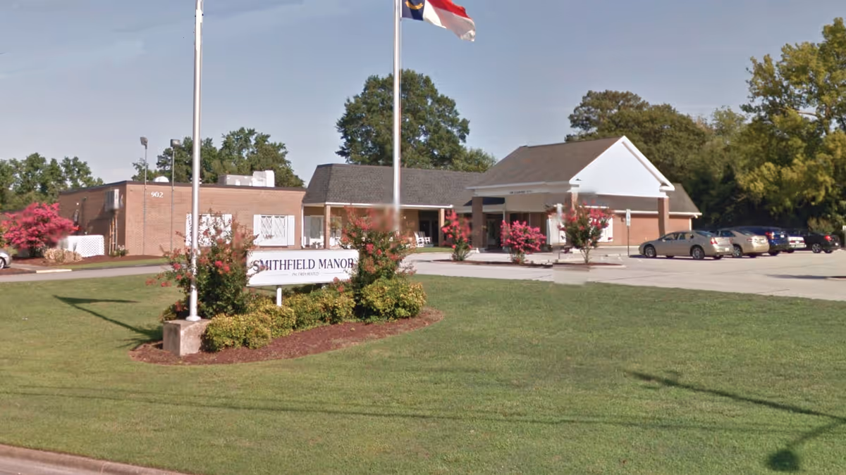 Front exterior of Smithfield Manor nursing facility showing the entrance canopy, flagpoles, landscaped sign, and parking lot.