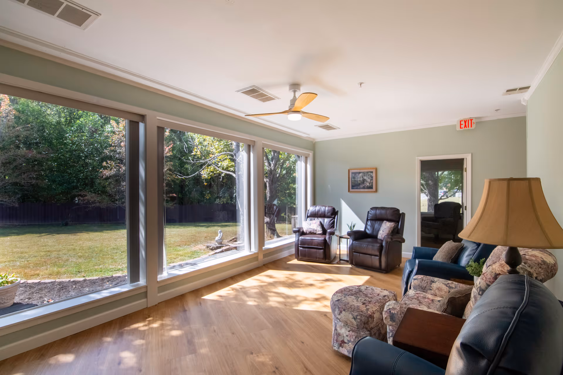 A bright and cozy living room with large windows overlooking a green outdoor area. The room features two dark leather recliners with pillows, a floral-patterned armchair with a matching ottoman, a blue armchair, a wooden side table, and a lamp with a beige shade. The walls are painted light green, and there is a ceiling fan with wooden blades. Sunlight streams through the windows, casting shadows on the wooden floor.