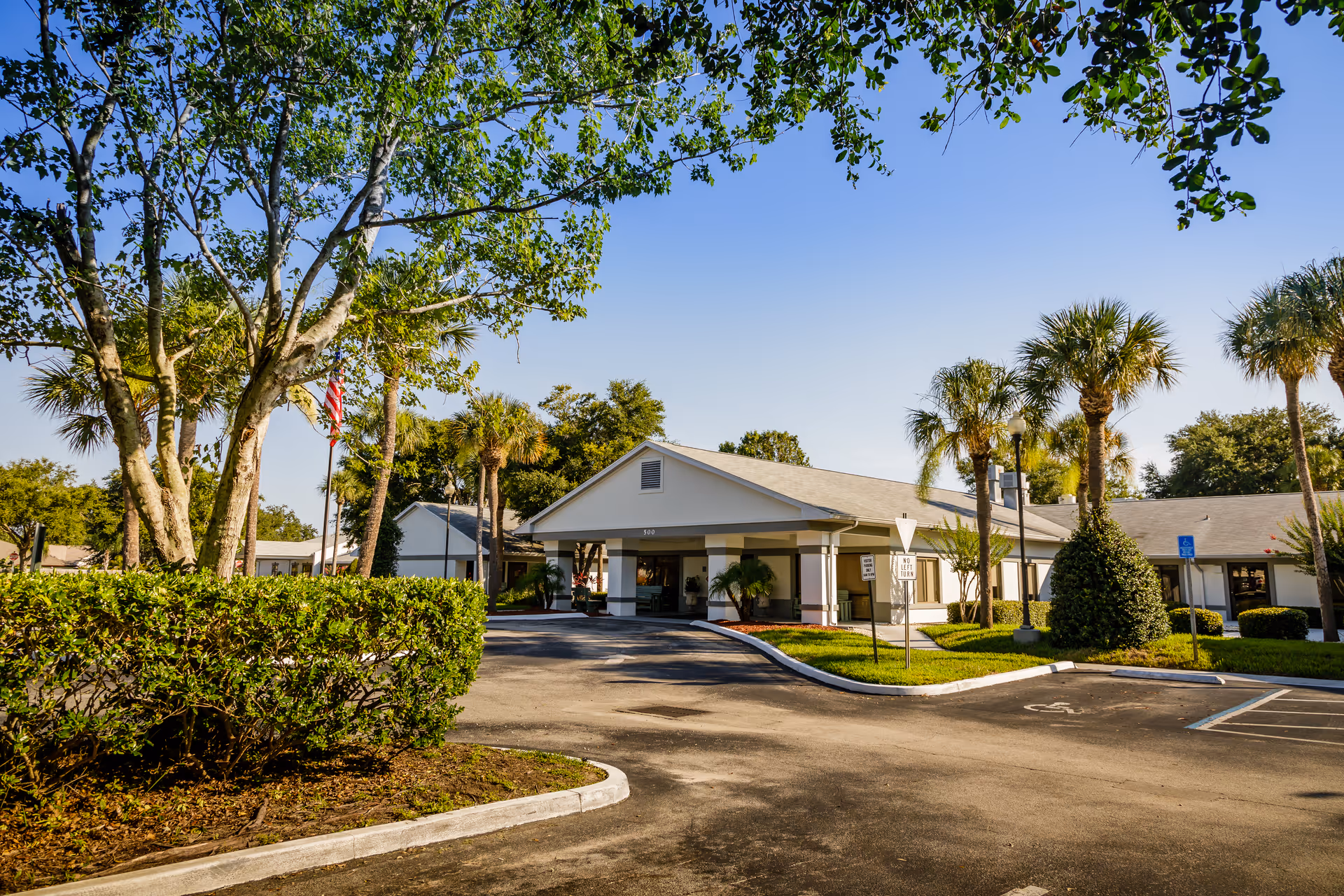 Sunlit senior care facility with a covered entrance, palm trees, and a paved driveway.