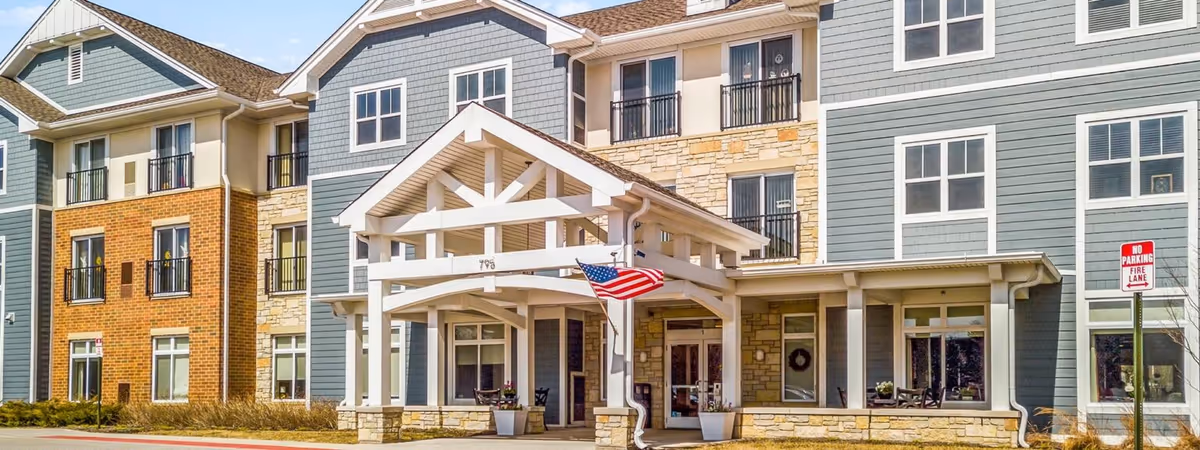 Exterior view of a senior living facility named Evolve at Lake Zurich featuring a multi-story building with a combination of blue siding, brick, and stone accents. The entrance has a white wooden pergola with an American flag hanging from it. There are several windows and small balconies visible, along with a 'No Parking Fire Lane' sign near the entrance.
